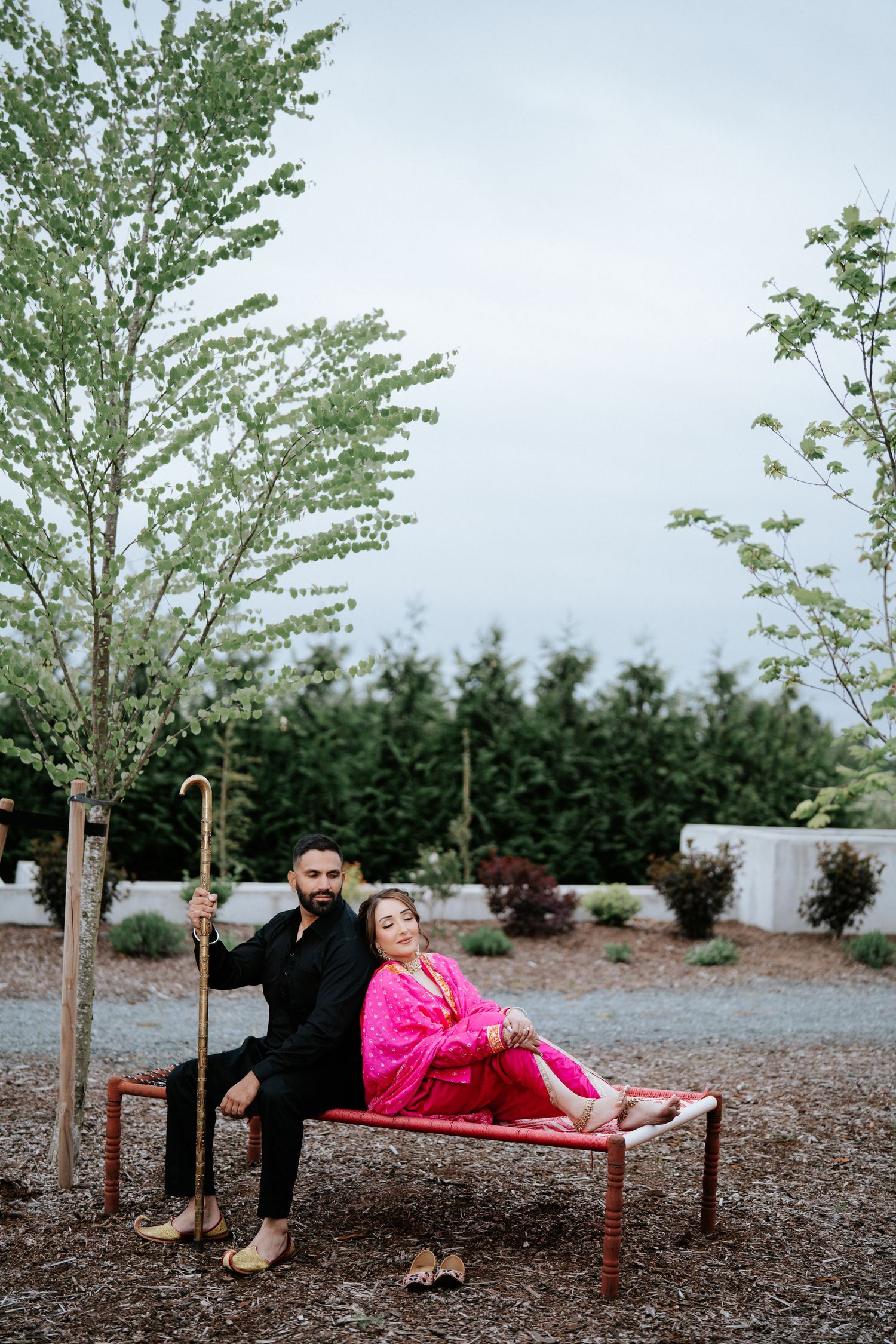 A couple sits on a red bench outdoors, man in black holding a cane, woman in bright pink outfit leaning against him.