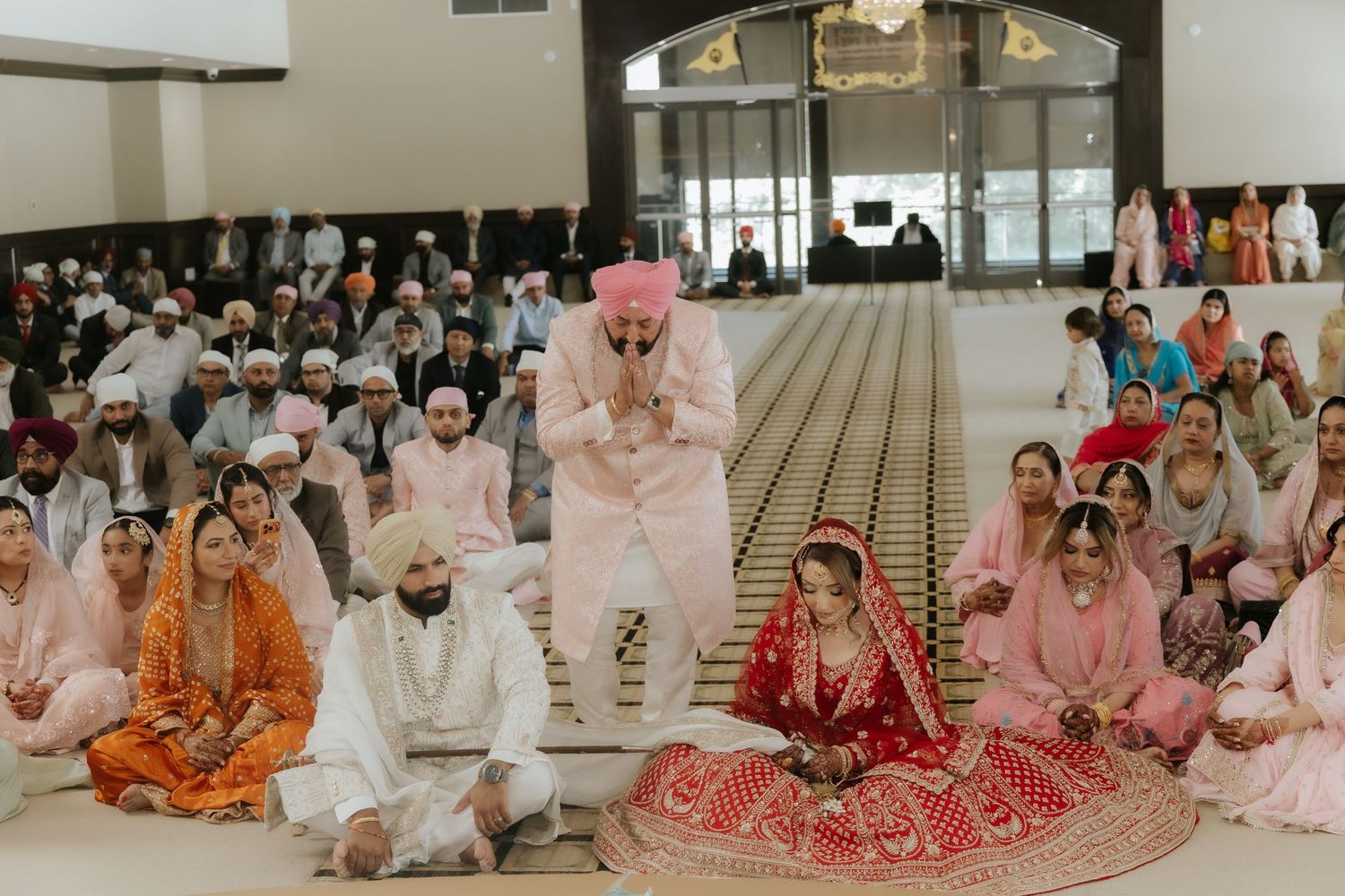 Sikh wedding ceremony with bride in red lehenga and groom in pink turban seated on floor in a gurudwara.
