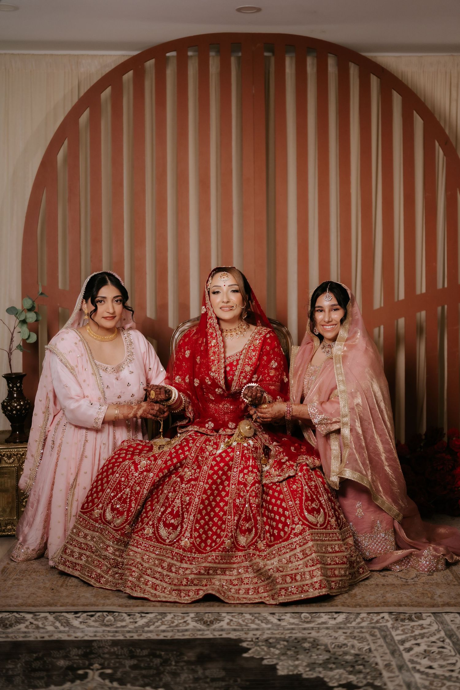Bride in red bridal lehenga seated with two women in pink outfits before an arch backdrop.