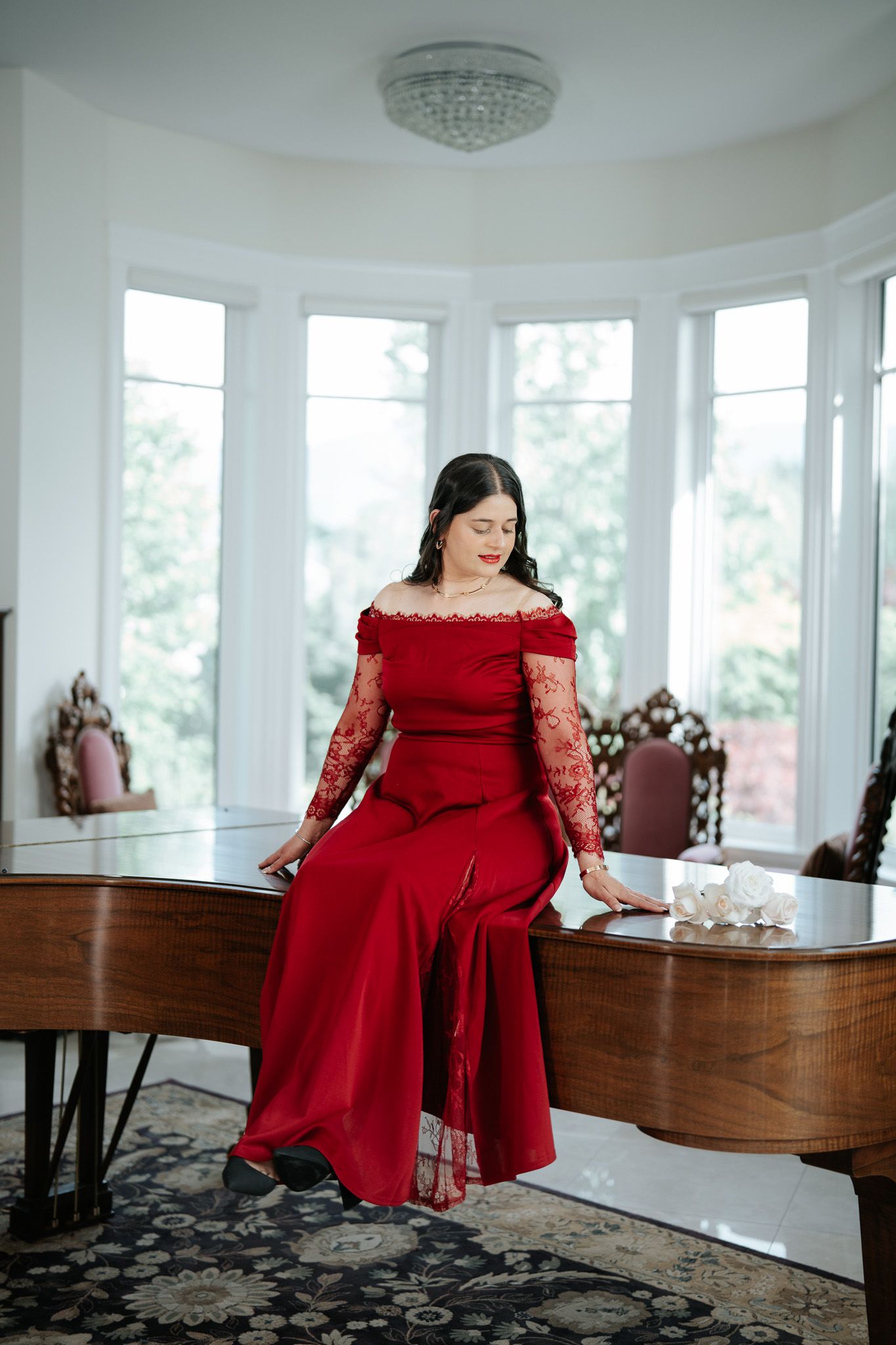 Woman in red off-shoulder gown sits gracefully on a grand piano by bay windows.