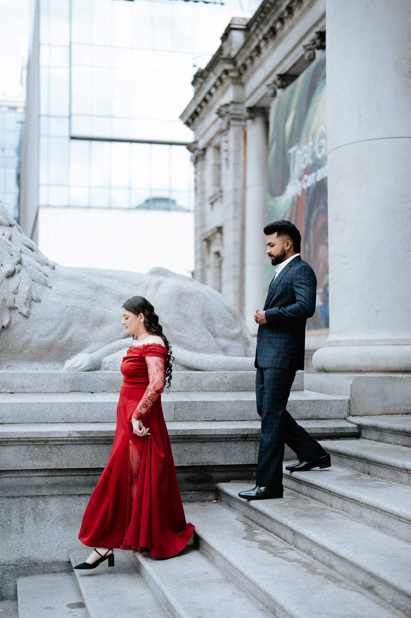 Elegant couple on museum steps, woman in red off-shoulder gown and man in dark suit, classic architecture backdrop.