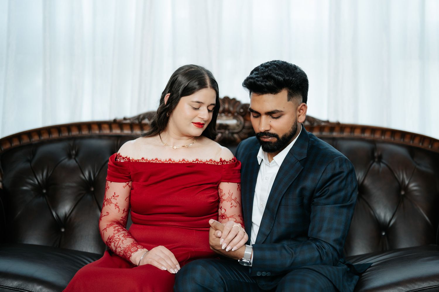 Couple in formal attire sitting together on leather sofa, woman in red lace dress.