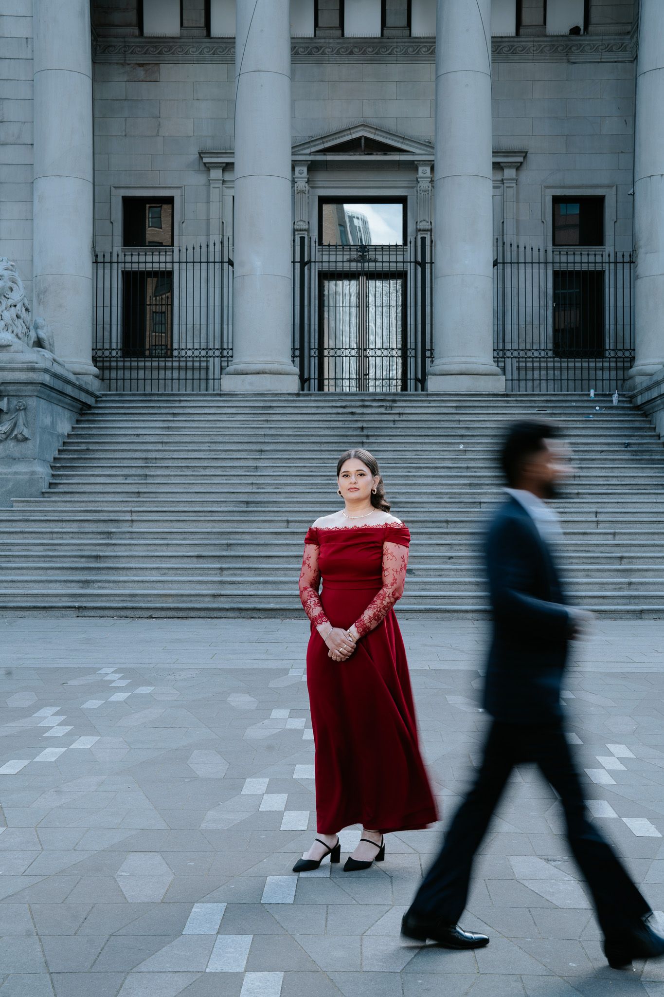 Woman in red gown stands still before grand building as blurred man in suit walks past.