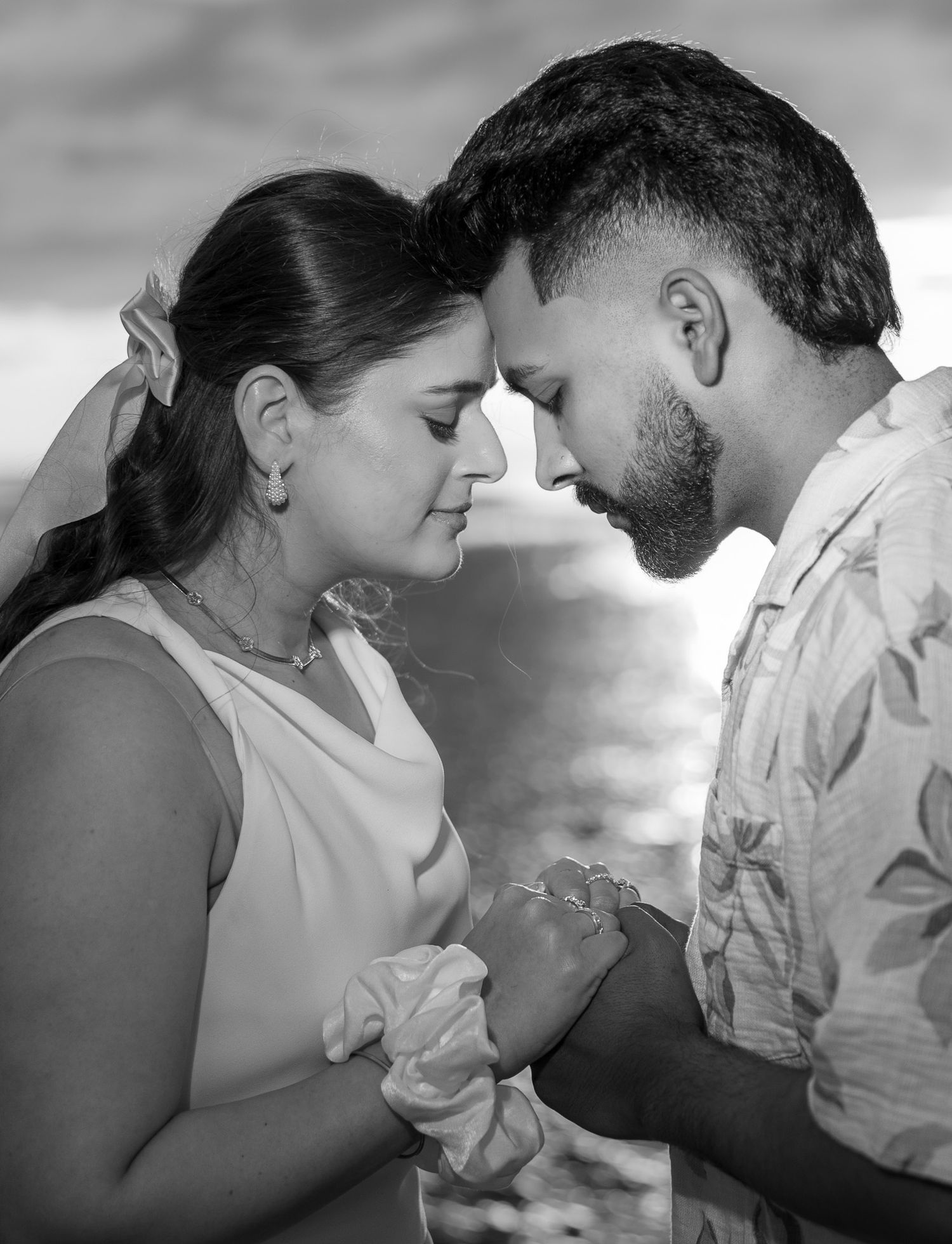 Black and white romantic couple touching foreheads tenderly while holding hands outdoors near ocean.