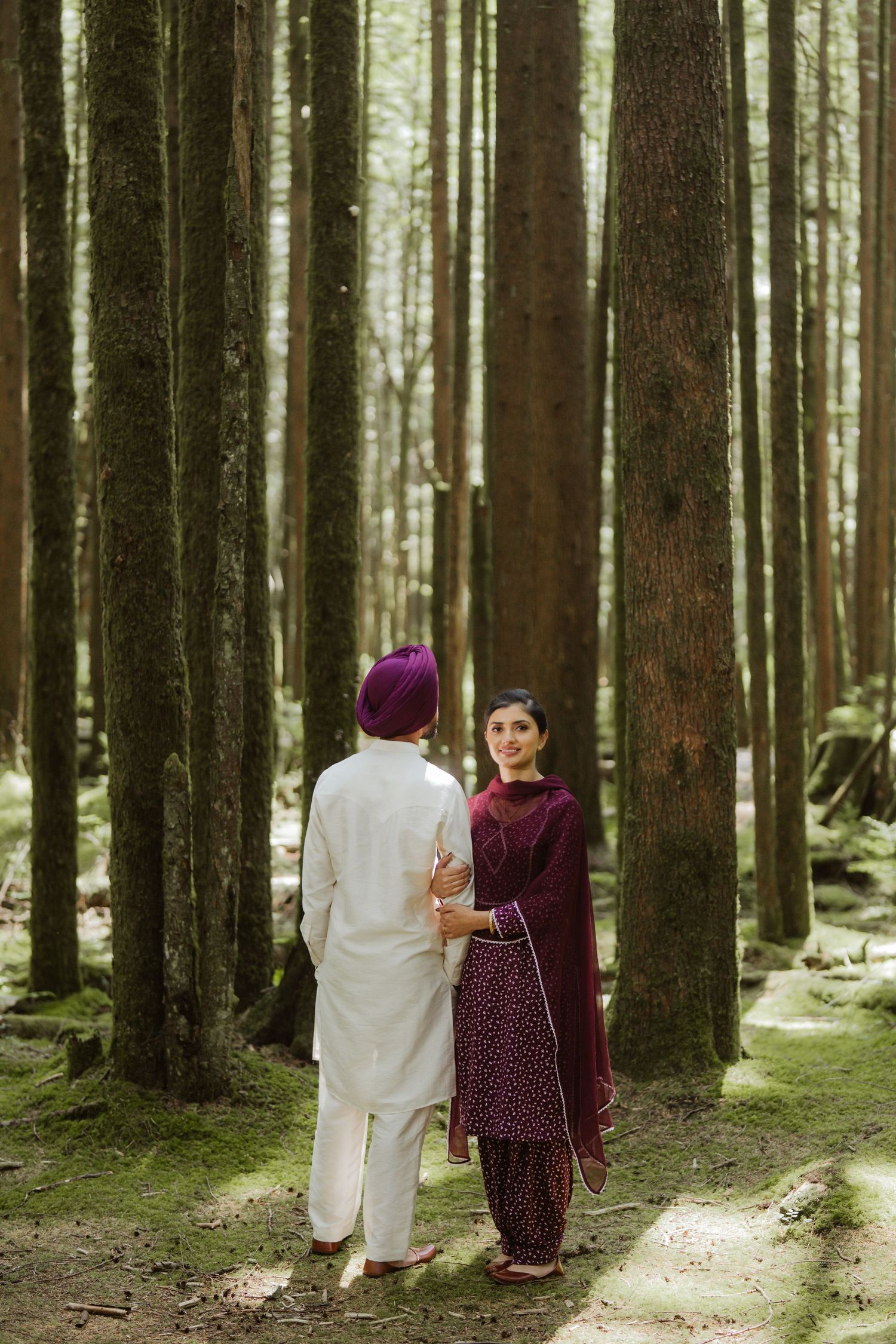 Sikh couple in traditional attire walking through a sunlit forest, man in white kurta and purple turban, woman in maroon salwar.