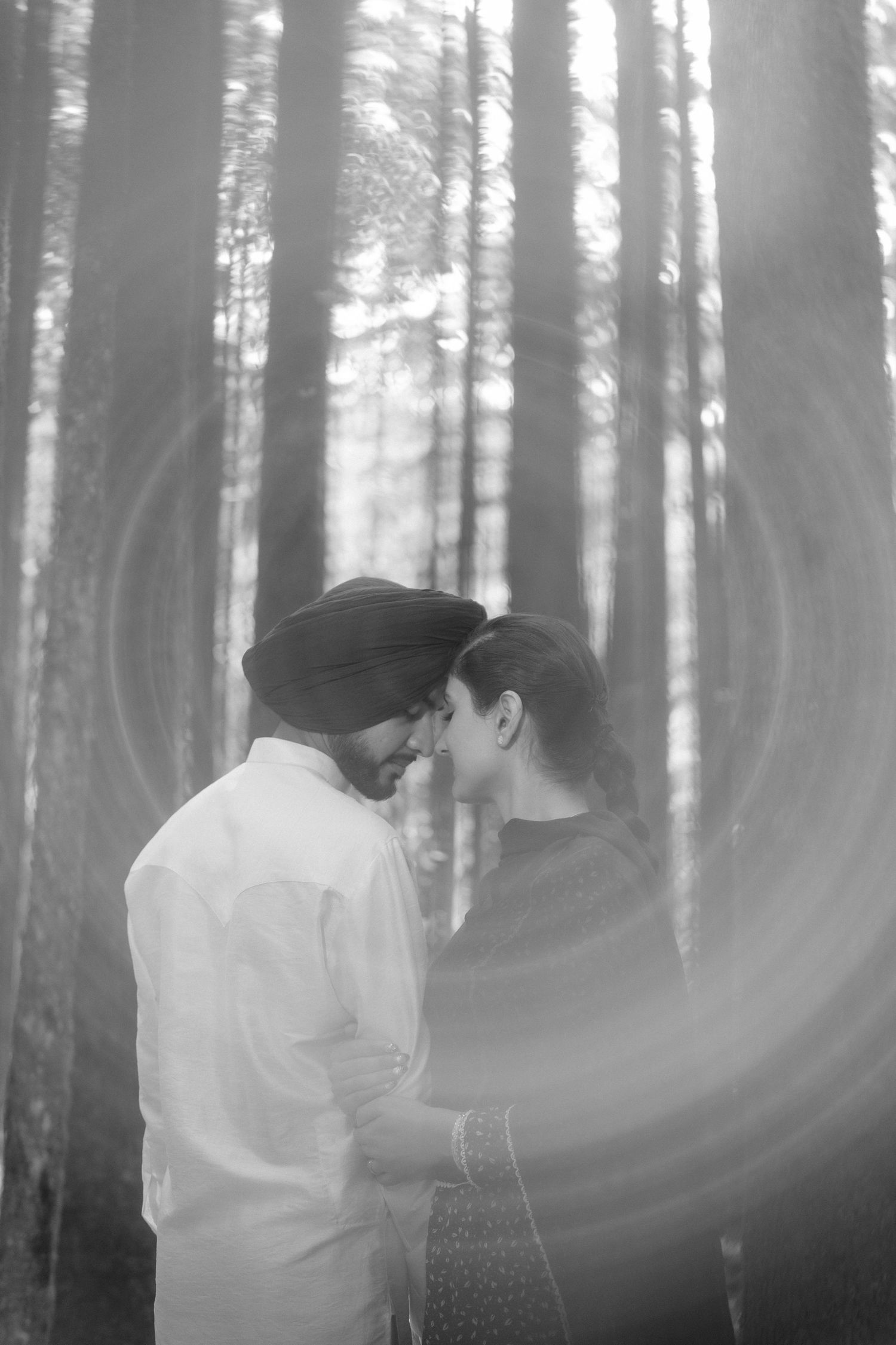 Black and white romantic portrait of a Sikh couple touching foreheads in a sunlit forest.