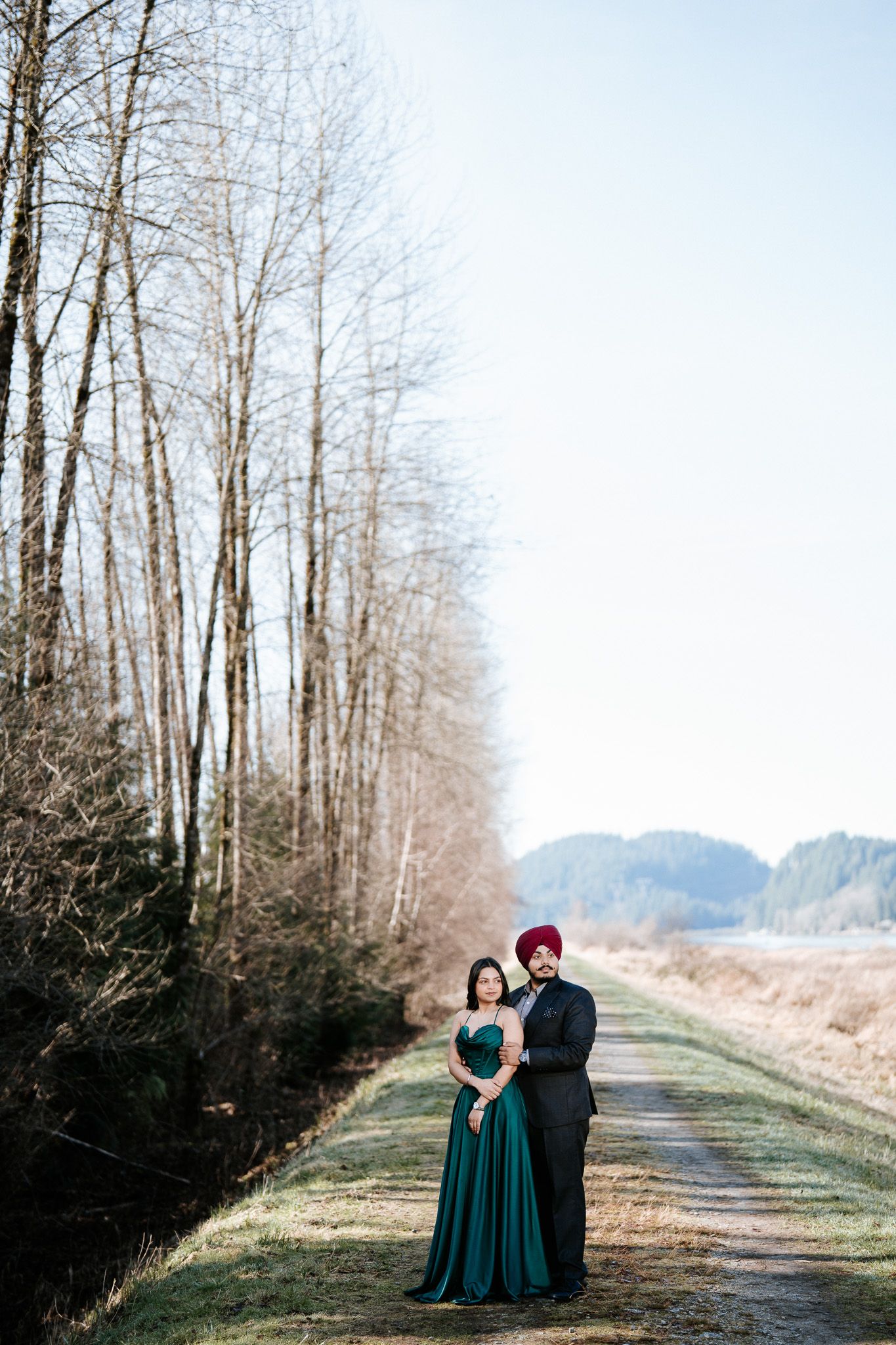 A couple in formal attire embraces on a tree-lined path with mountains visible in the background.