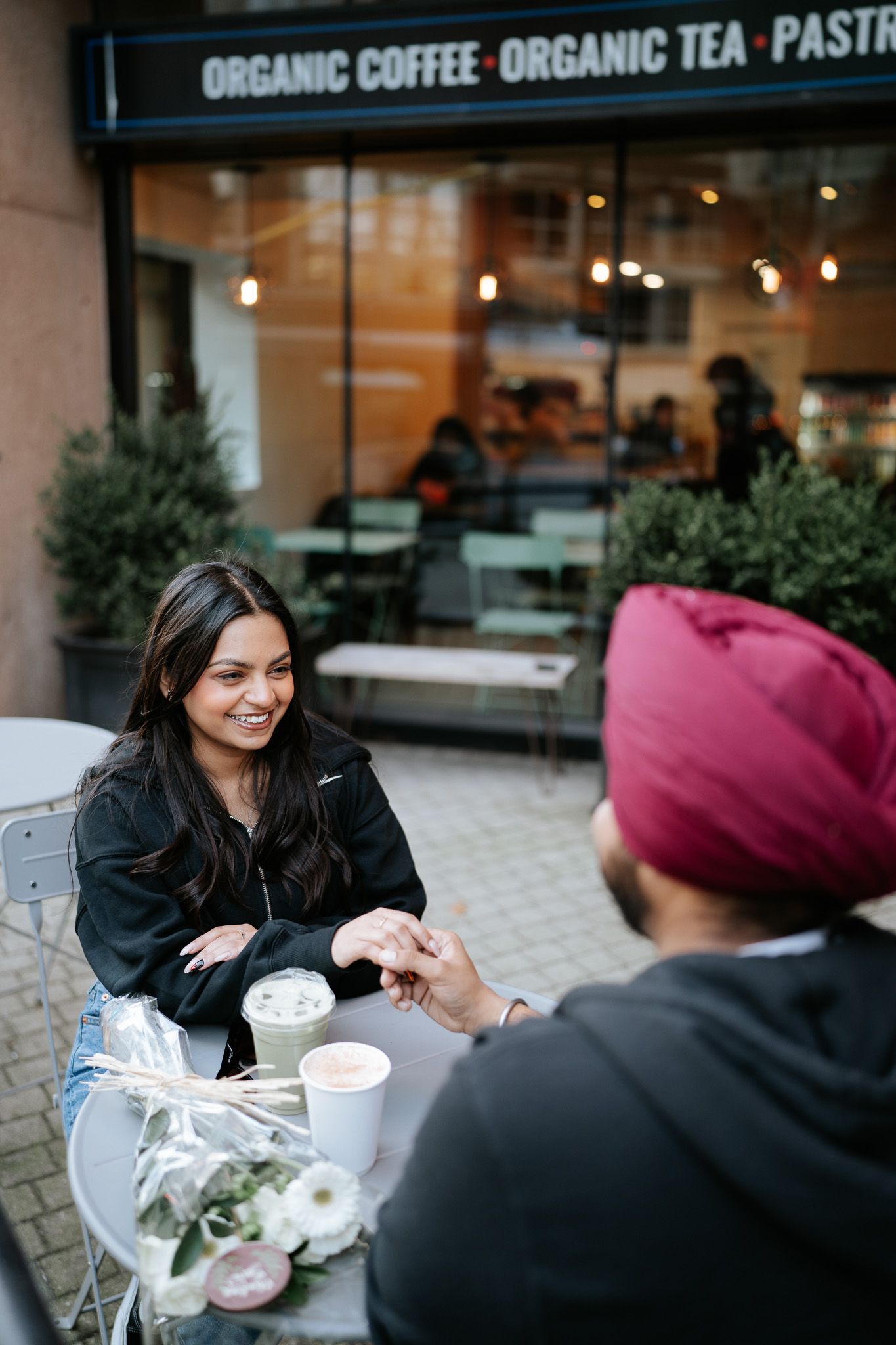 Two people having coffee at an outdoor cafe table with organic coffee and tea signage visible in background.