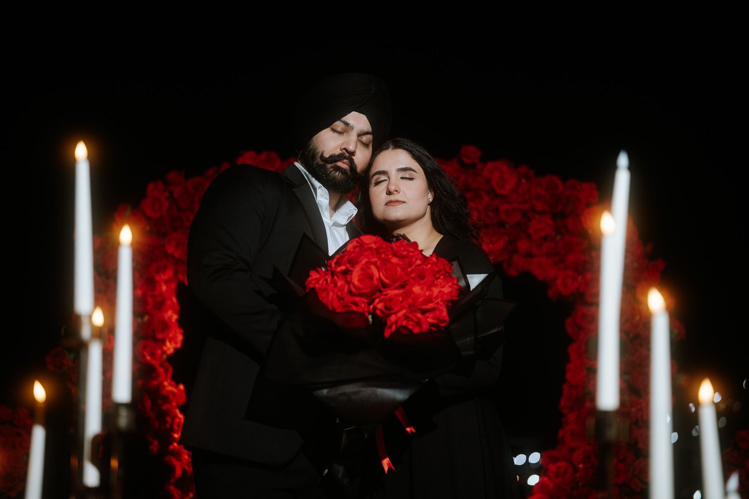 Romantic couple embraces near candlelight and red roses in a dark, moody setting.