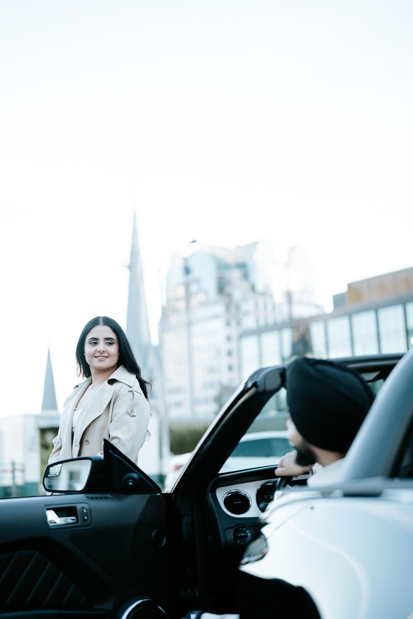 A person stands by a luxury car in an urban setting with modern buildings in the background.