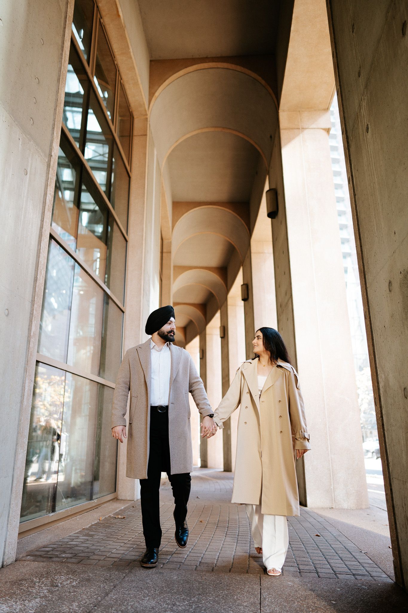 A couple in matching beige coats walks hand in hand through an elegant arched colonnade hallway.
