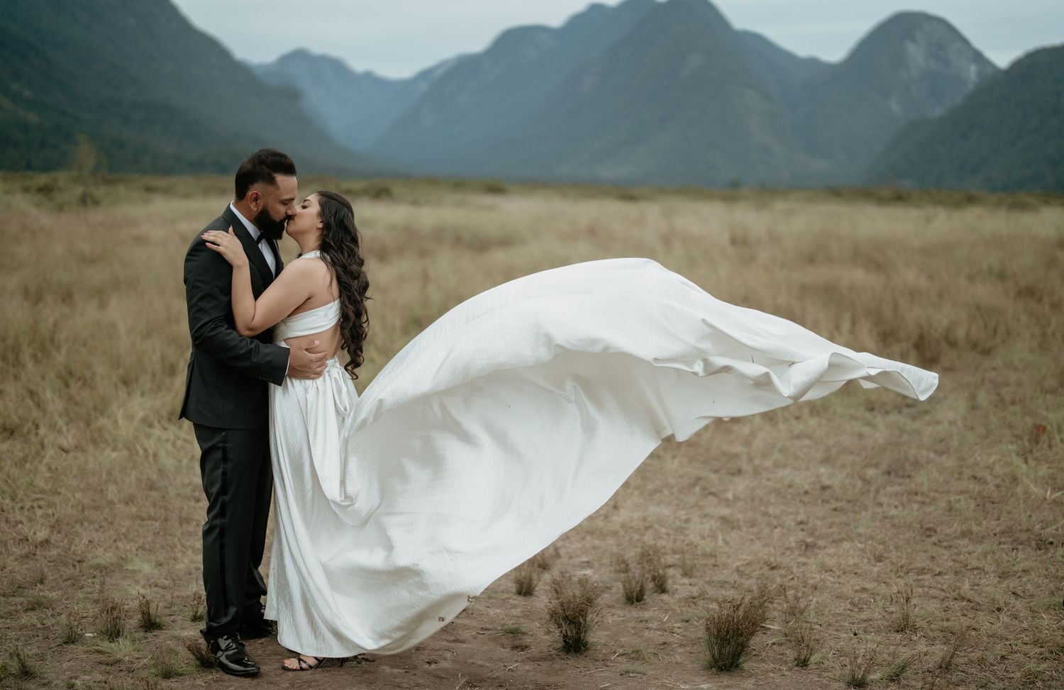 A romantic wedding photo showing a flowing white dress billowing in the wind against a dramatic mountain landscape.