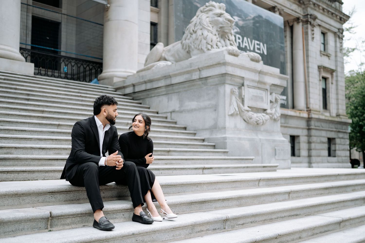 A couple sits together on white stone steps of a grand building with columns.