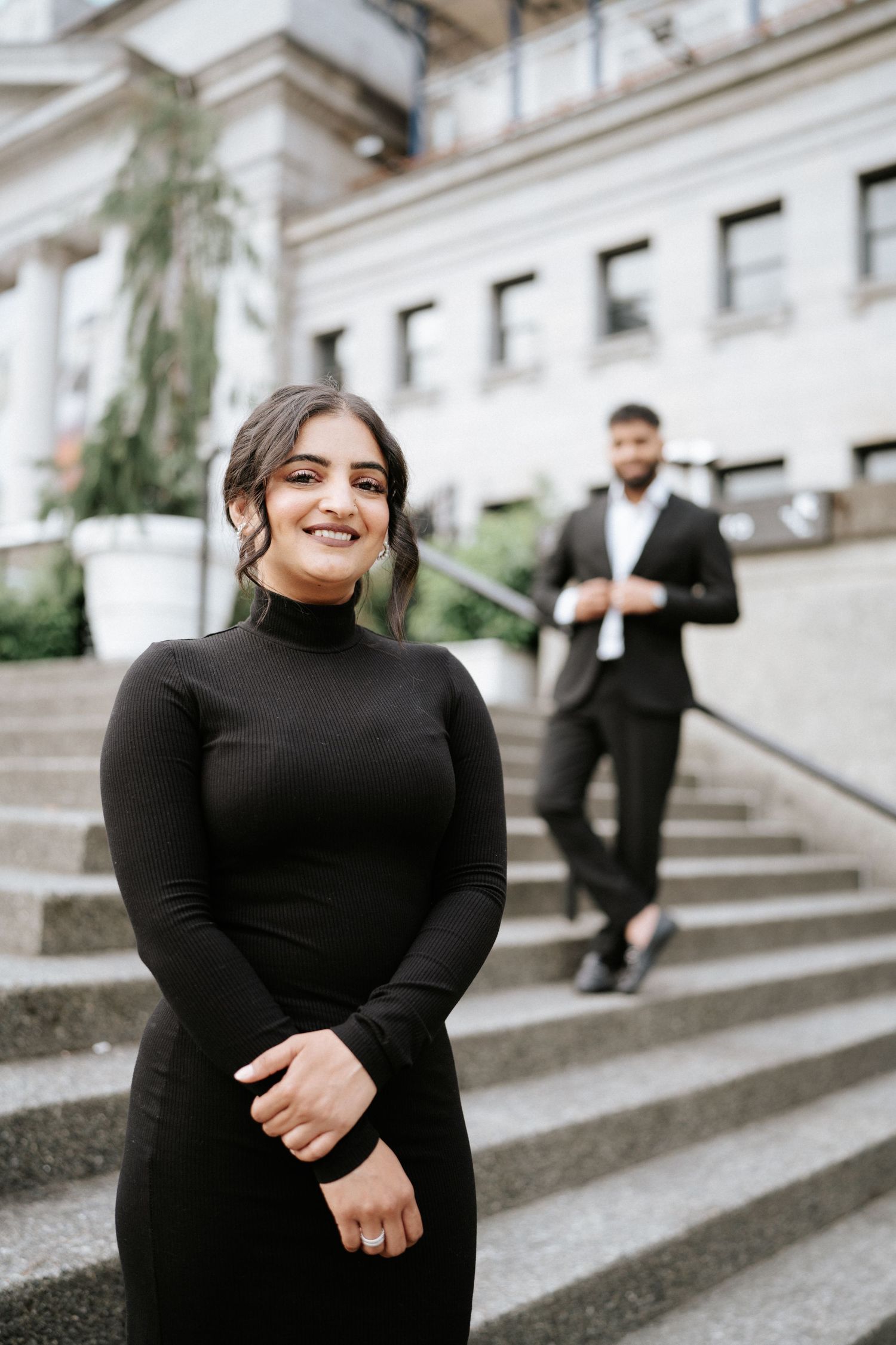 A couple in elegant black attire poses on the steps of a classical building with white columns.