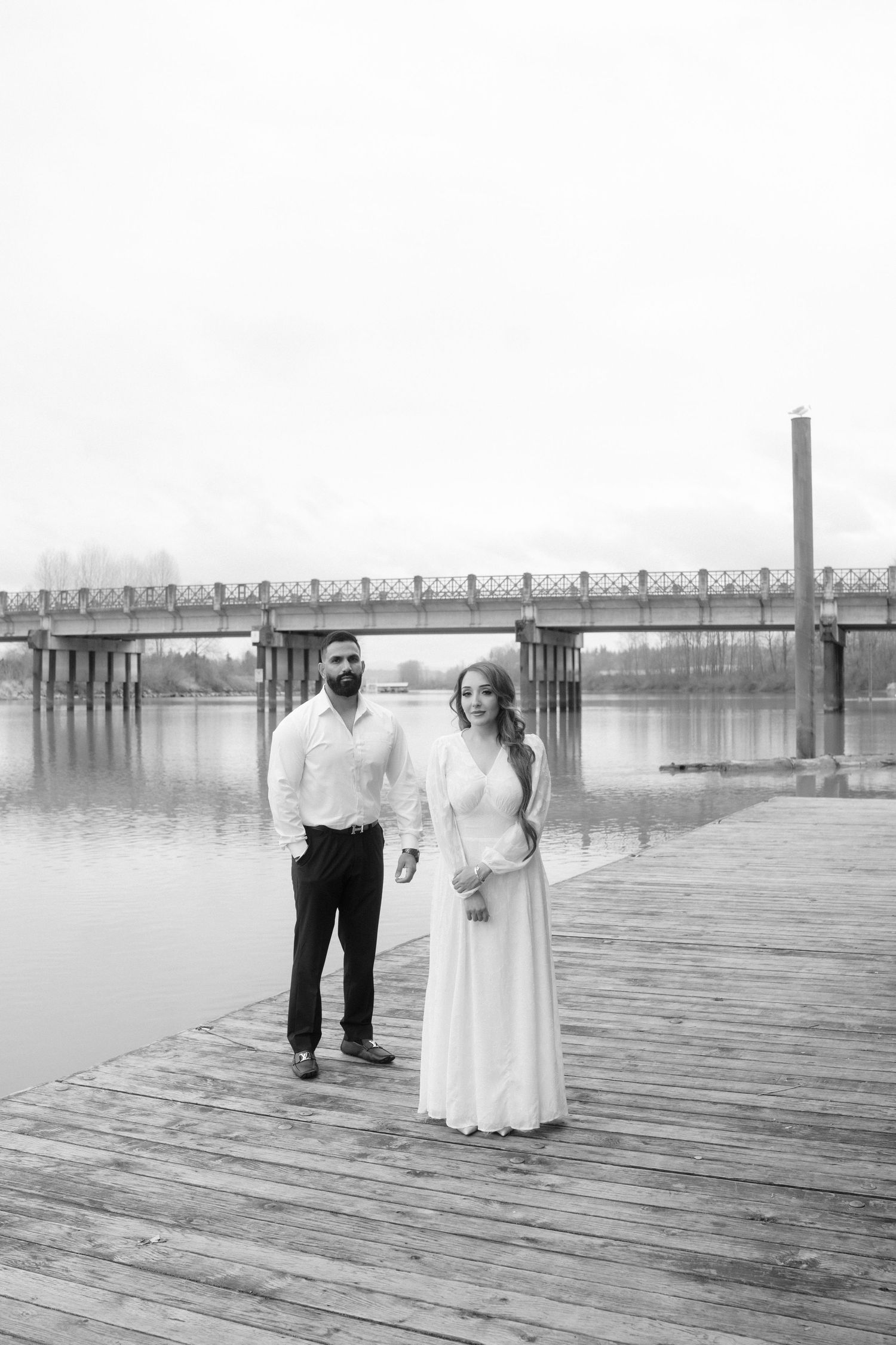 A couple stands together on a wooden pier with a bridge visible in the foggy distance.
