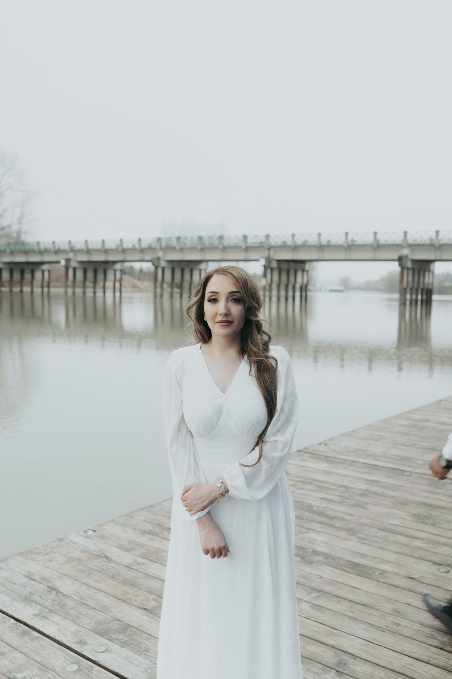 A woman in a flowing white dress poses elegantly on a wooden dock with a bridge behind her.