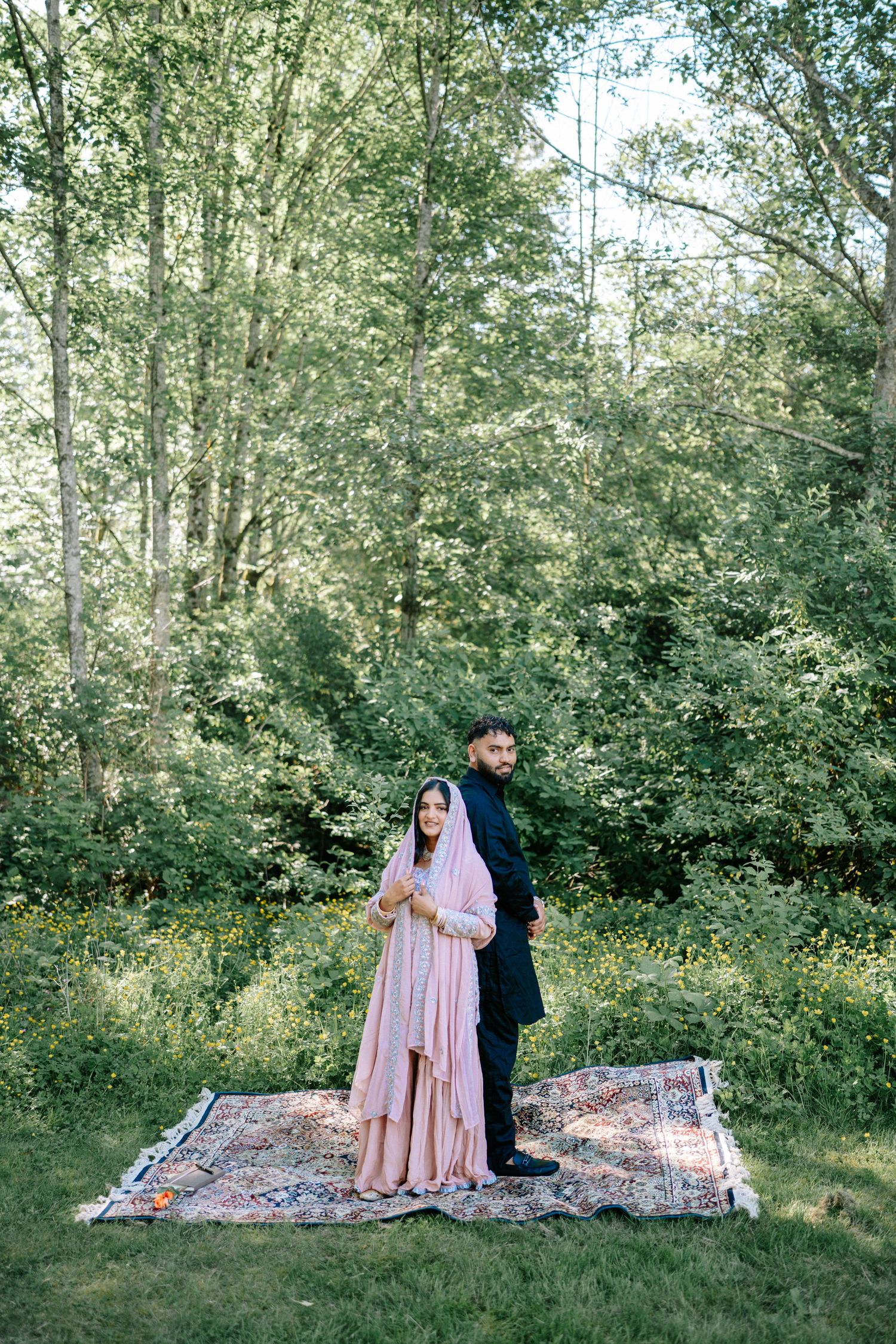 A romantic couple poses on a picnic blanket surrounded by green foliage and natural sunlight.