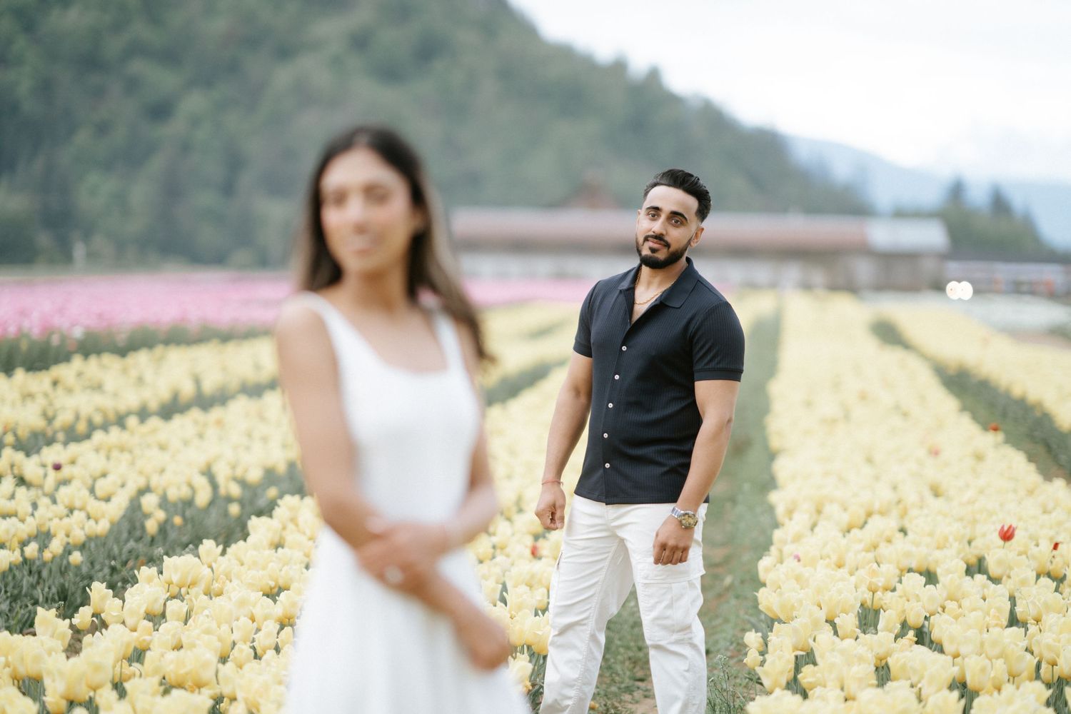 Two people pose in a scenic tulip field with rows of yellow flowers stretching into the distance near mountains.