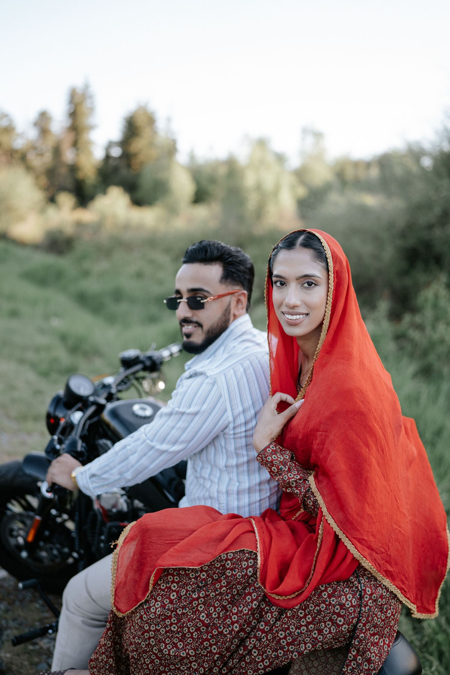 Couple on a motorcycle in traditional South Asian attire with a vibrant red dupatta flowing in the outdoor setting.