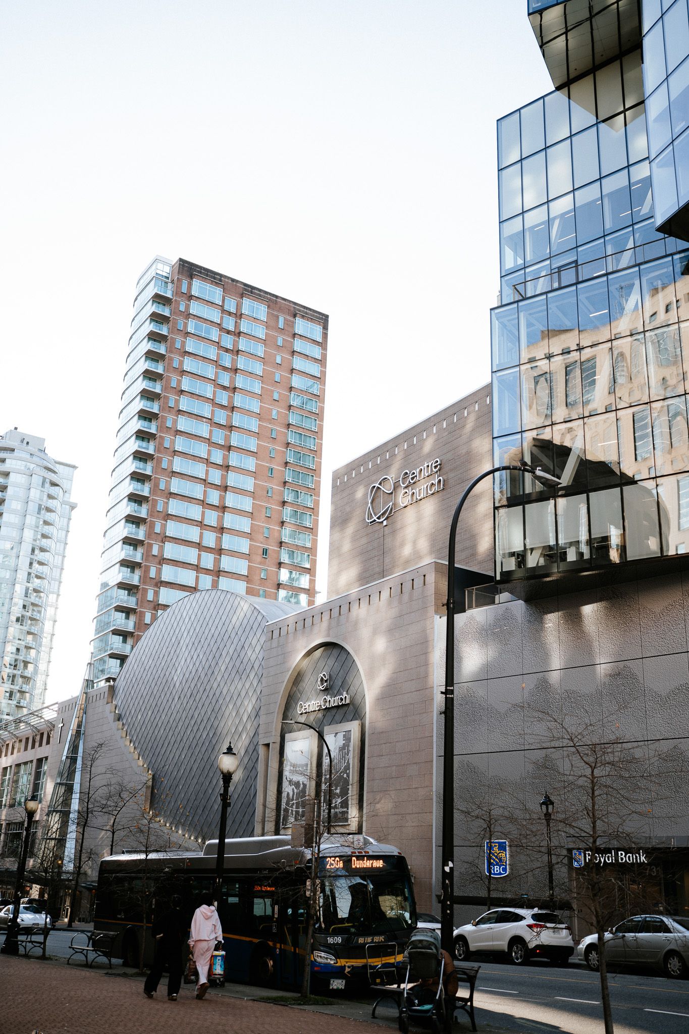 Modern glass and steel buildings reflect sunlight on a downtown street corner with parked cars and pedestrians.