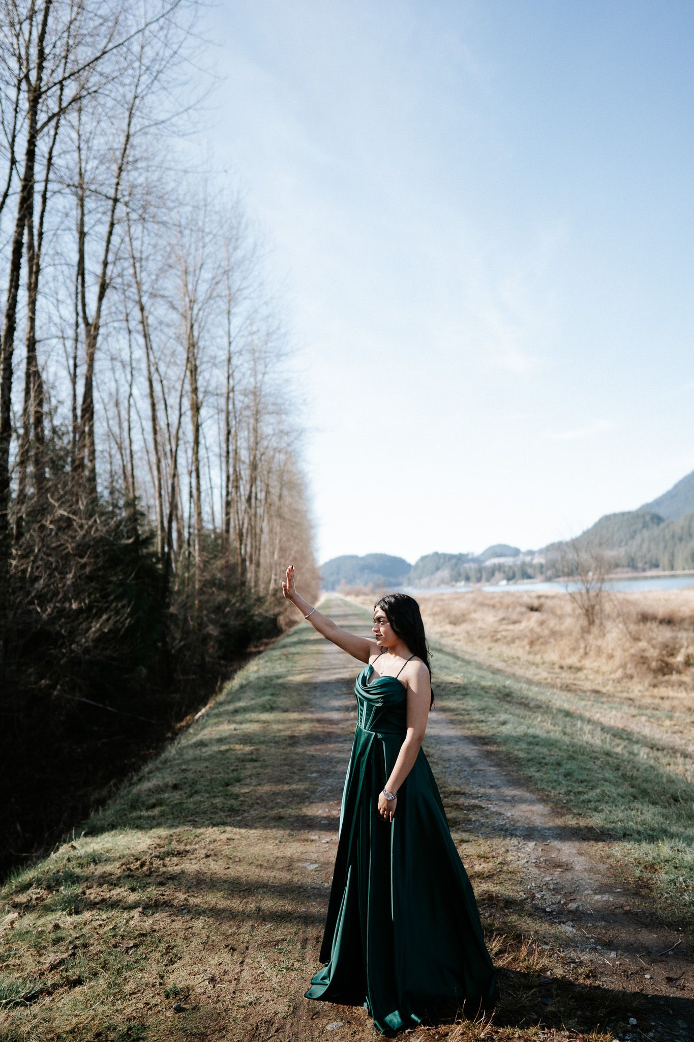 A person in an elegant emerald green gown stands on a scenic mountain trail surrounded by bare trees and mountains.