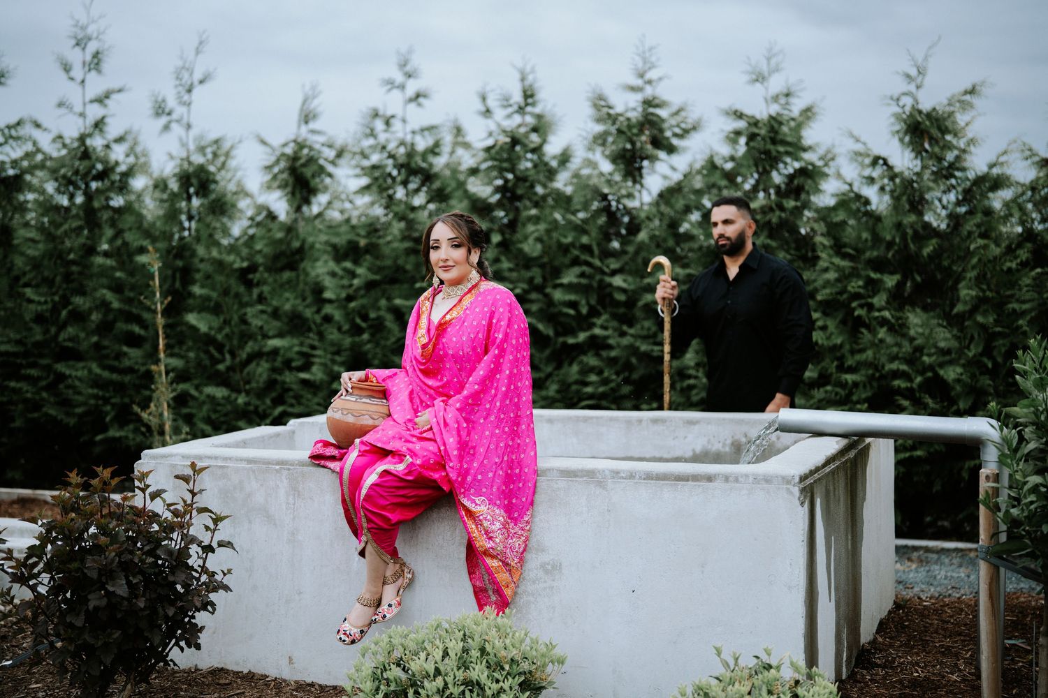 Woman in pink salwar kameez sits on white wall at tree farm while man in black stands behind her.