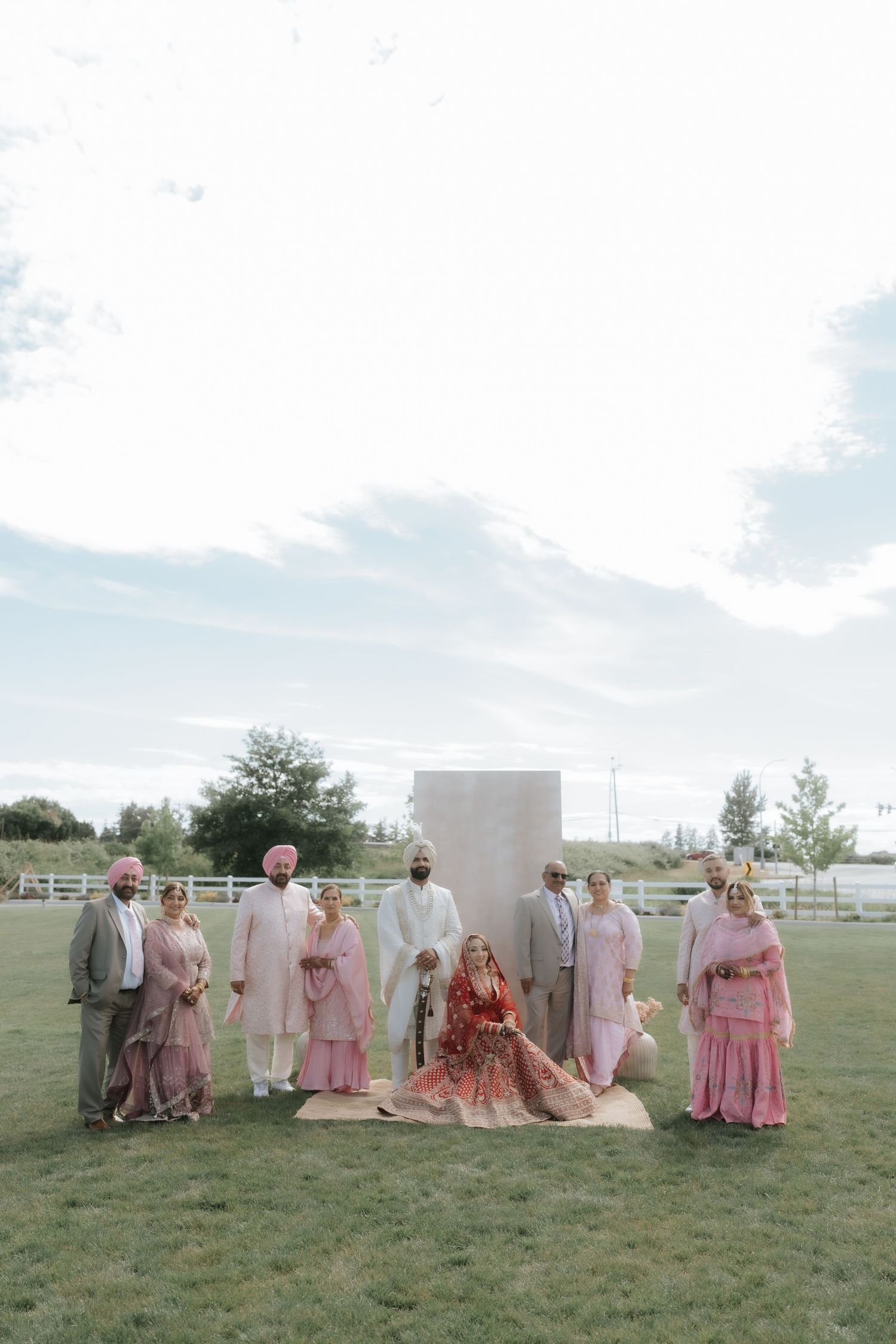 South Asian wedding party in pink and white attire poses outdoors on grass before a minimalist backdrop.