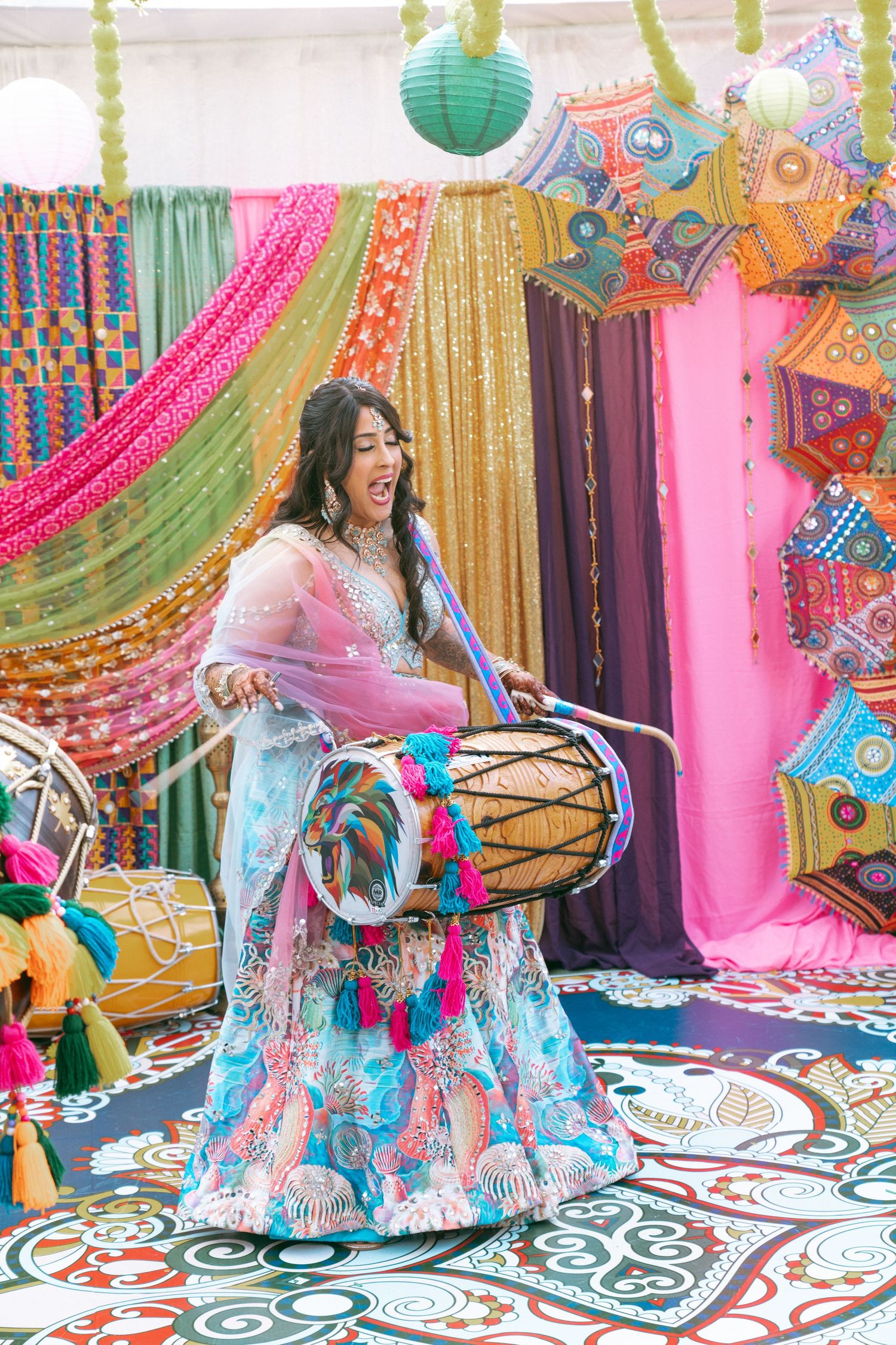 Woman in blue lehenga playing dhol drum at vibrant Indian wedding celebration with colorful decor.