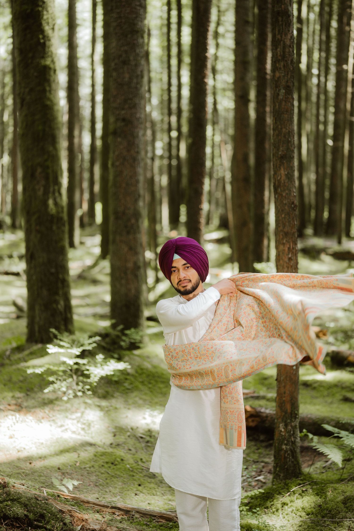 A Sikh man in white kurta and purple turban holds a shawl while standing in a lush green forest.