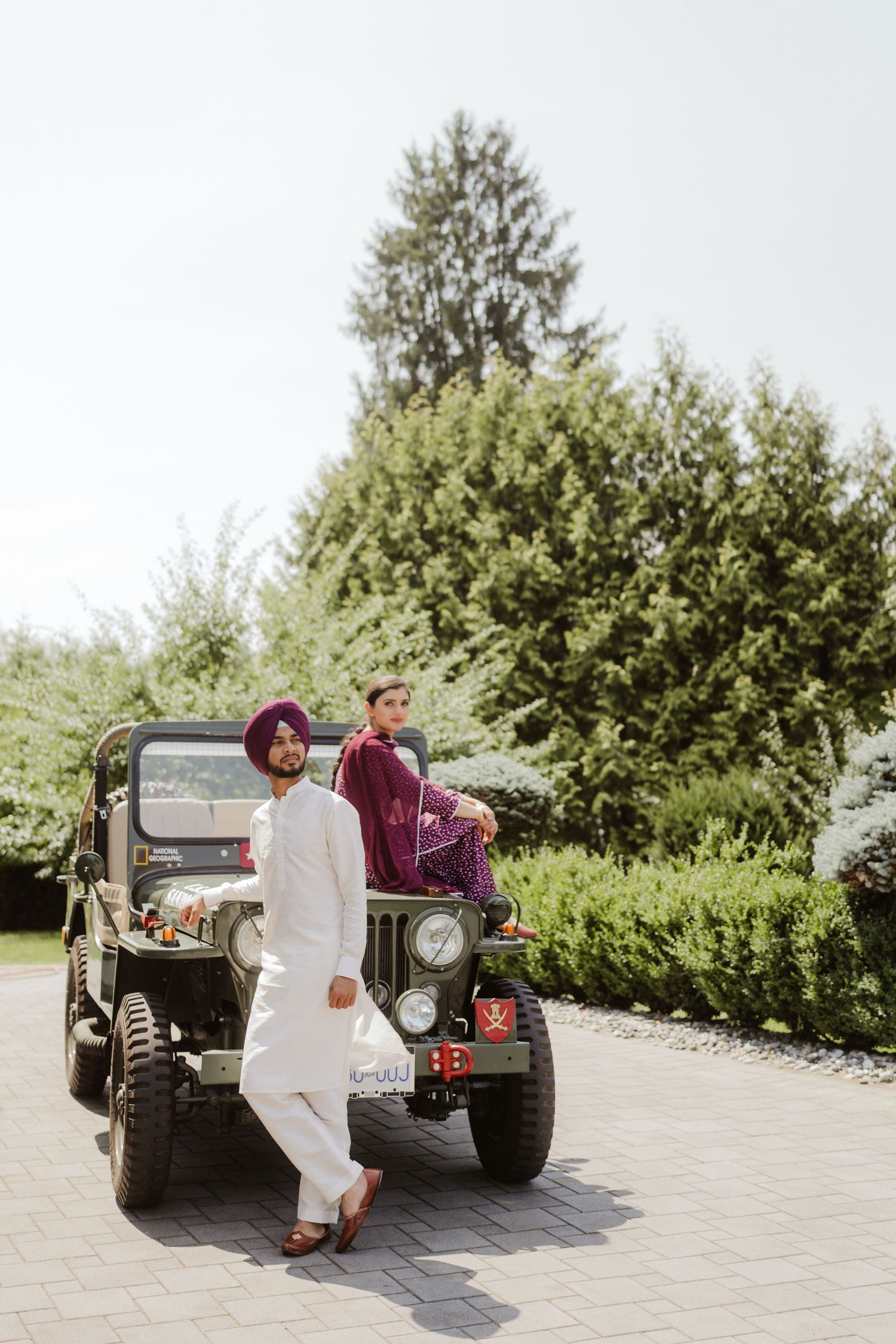 Sikh couple posing with a vintage military jeep outdoors, man in white kurta and woman in burgundy salwar kameez.