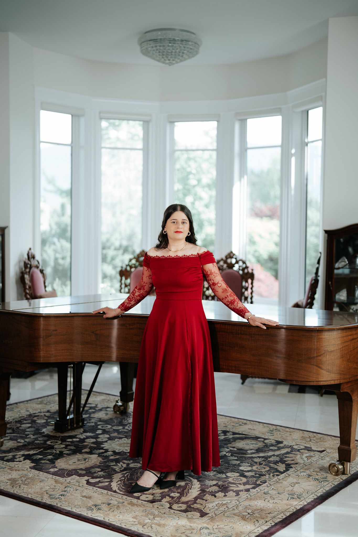 Woman in red off-shoulder ball gown standing before a grand piano in a bright elegant room.