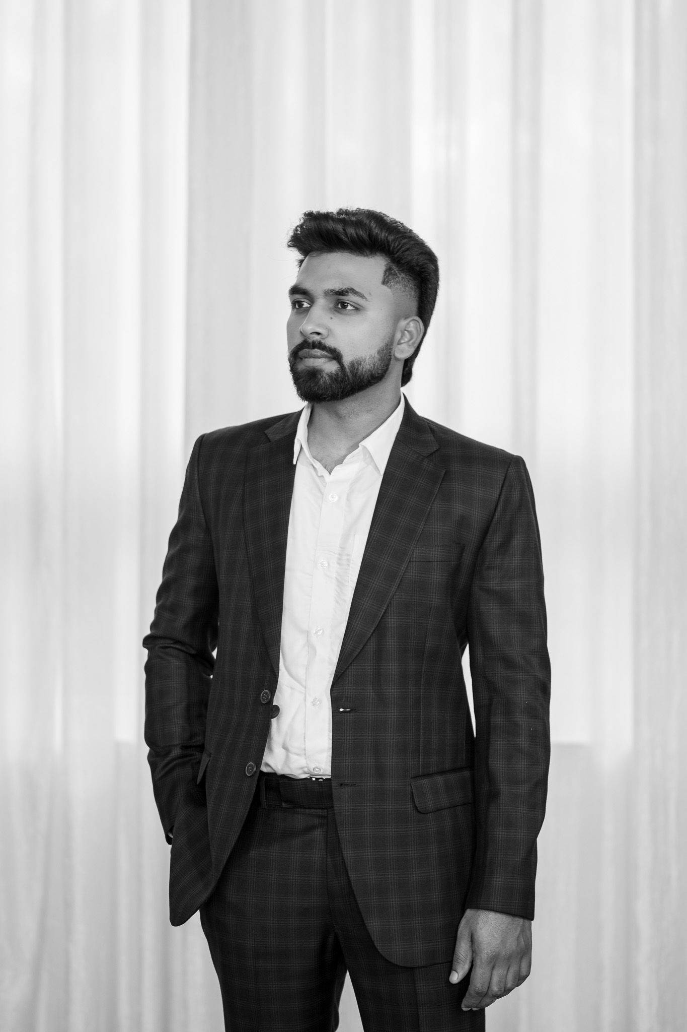 Stylish bearded man in plaid suit posing confidently against white curtain backdrop, black and white photo.