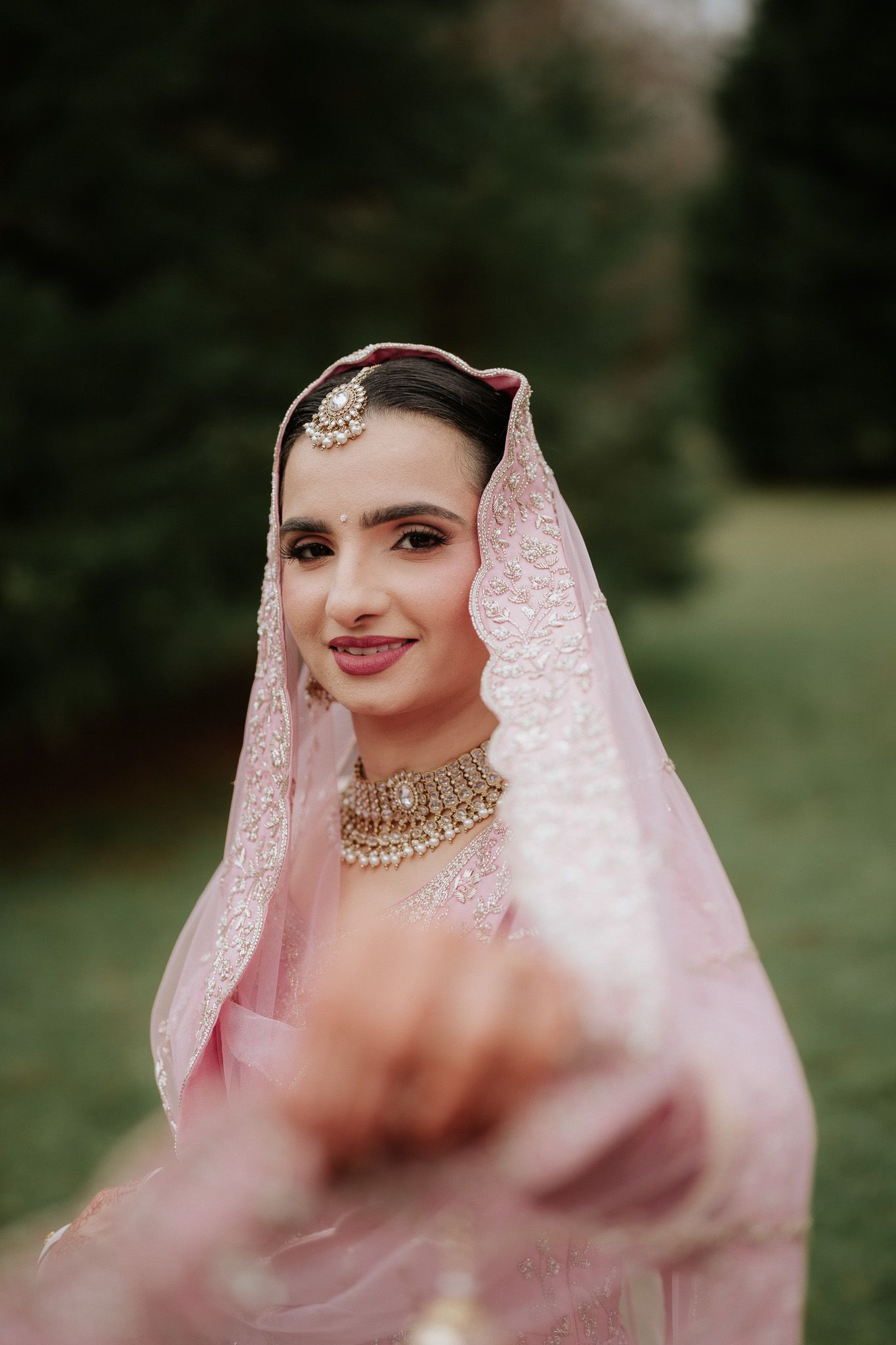 Close-up portrait of bride wearing pink traditional attire with ornate jewelry and delicate veil against natural backdrop.