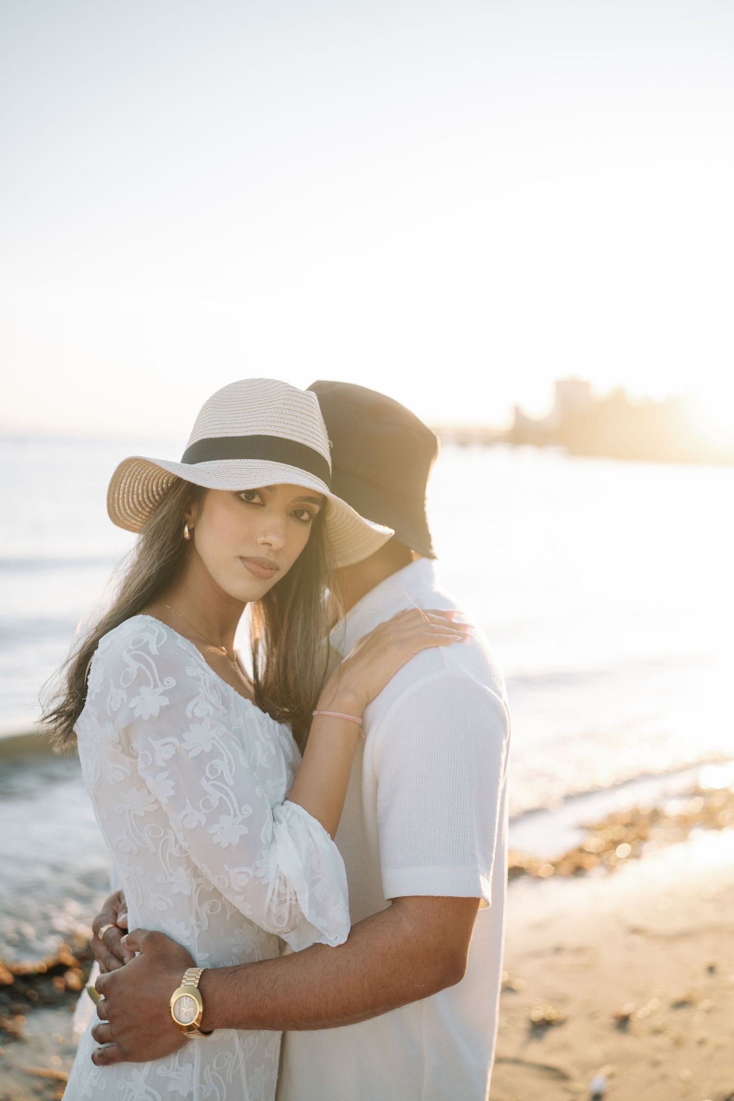 A couple embraces on a sunny beach during sunset wearing white outfits and a sun hat.