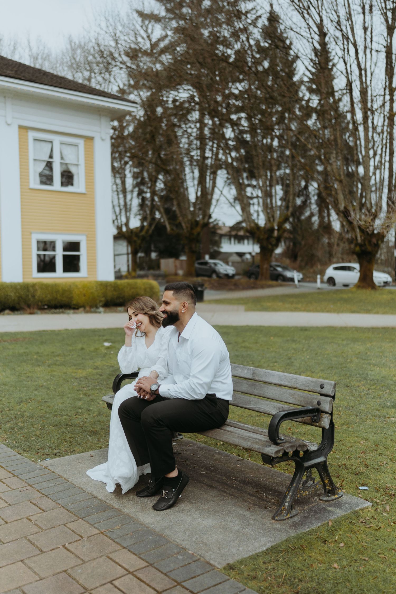 A couple shares a tender moment together on a wooden bench in a park setting with a yellow building in the background.