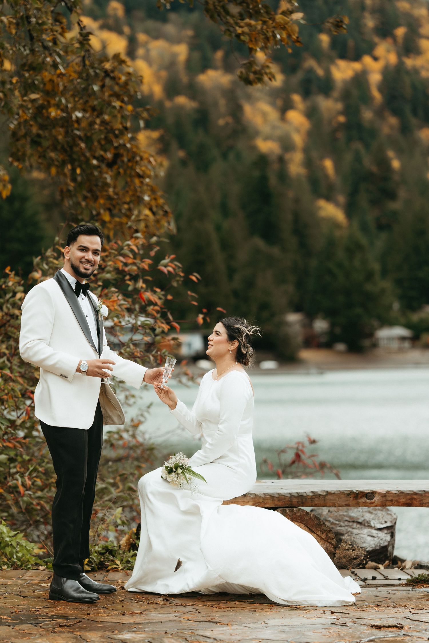 A couple in wedding attire playfully tosses autumn leaves by a lakeside during their fall wedding photoshoot.
