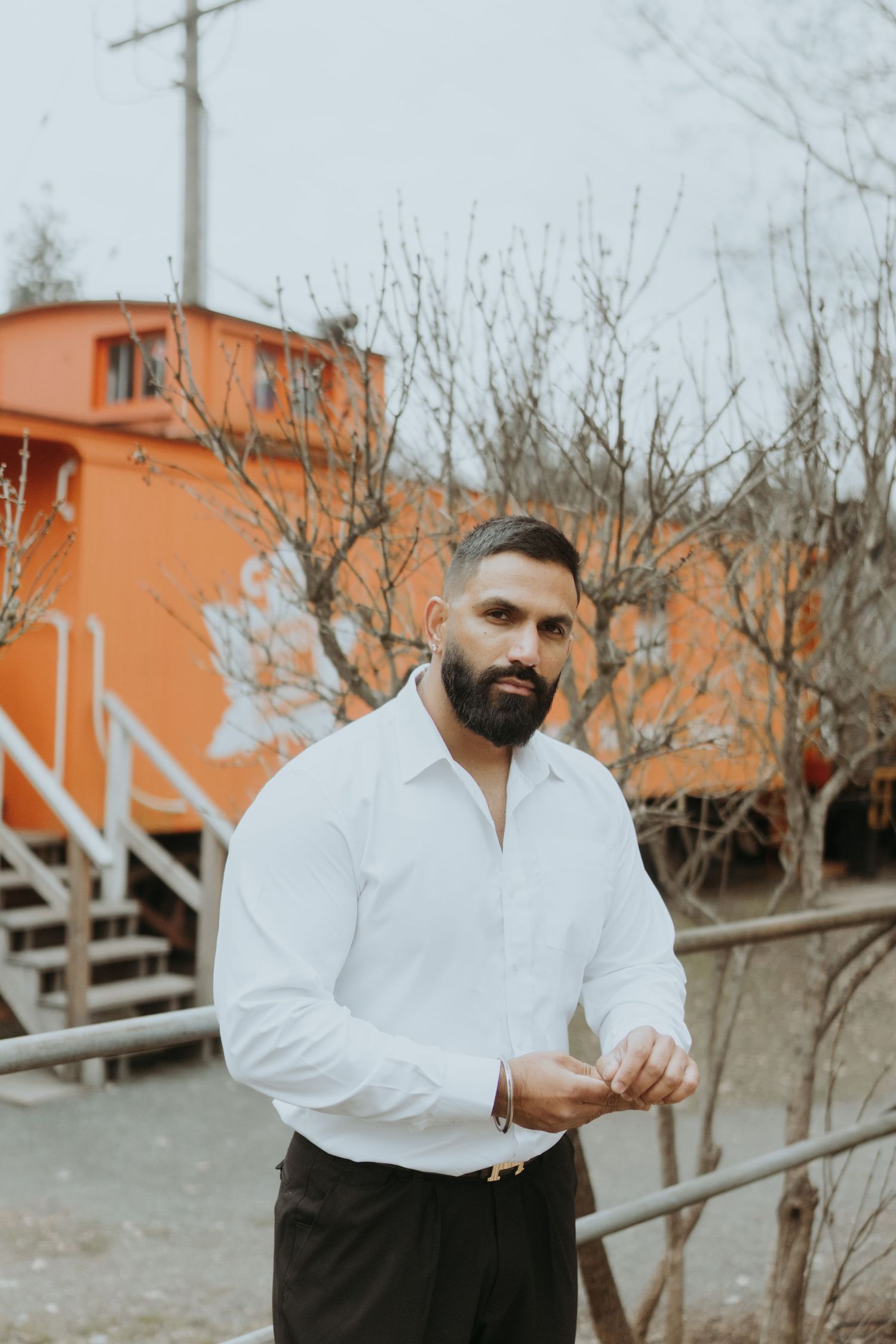 A person in formal attire gestures while standing near trees and an orange building.