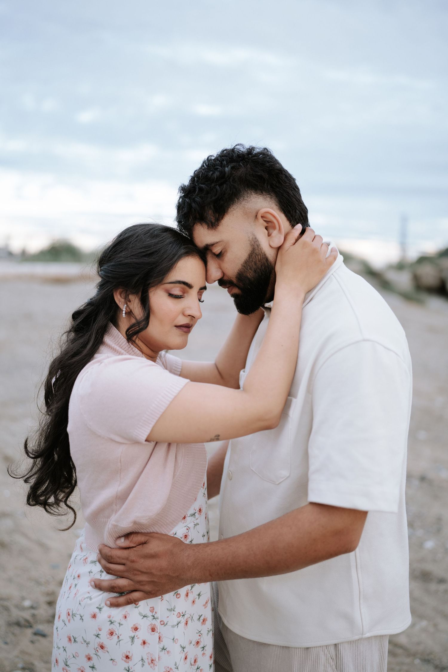 A couple in white and floral outfits shares a close embrace on the beach.
