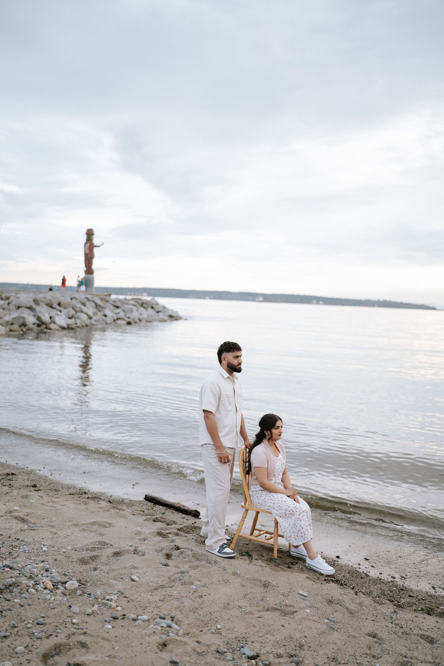 A couple in white outfits sits together on a rocky beach with mountains and water in the background.