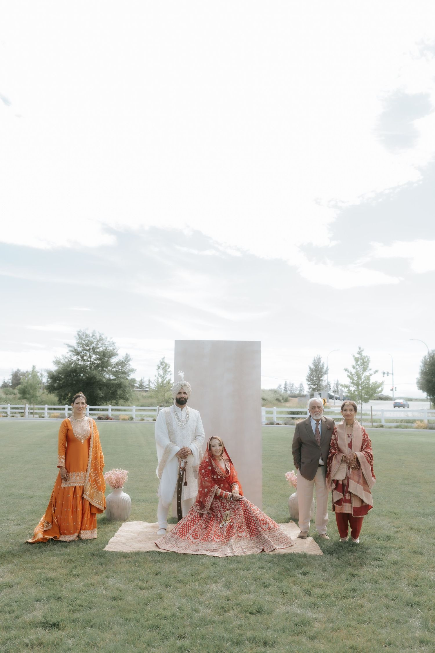 South Asian wedding party poses outdoors in traditional attire, bride seated in red lehenga with groom in white sherwani.