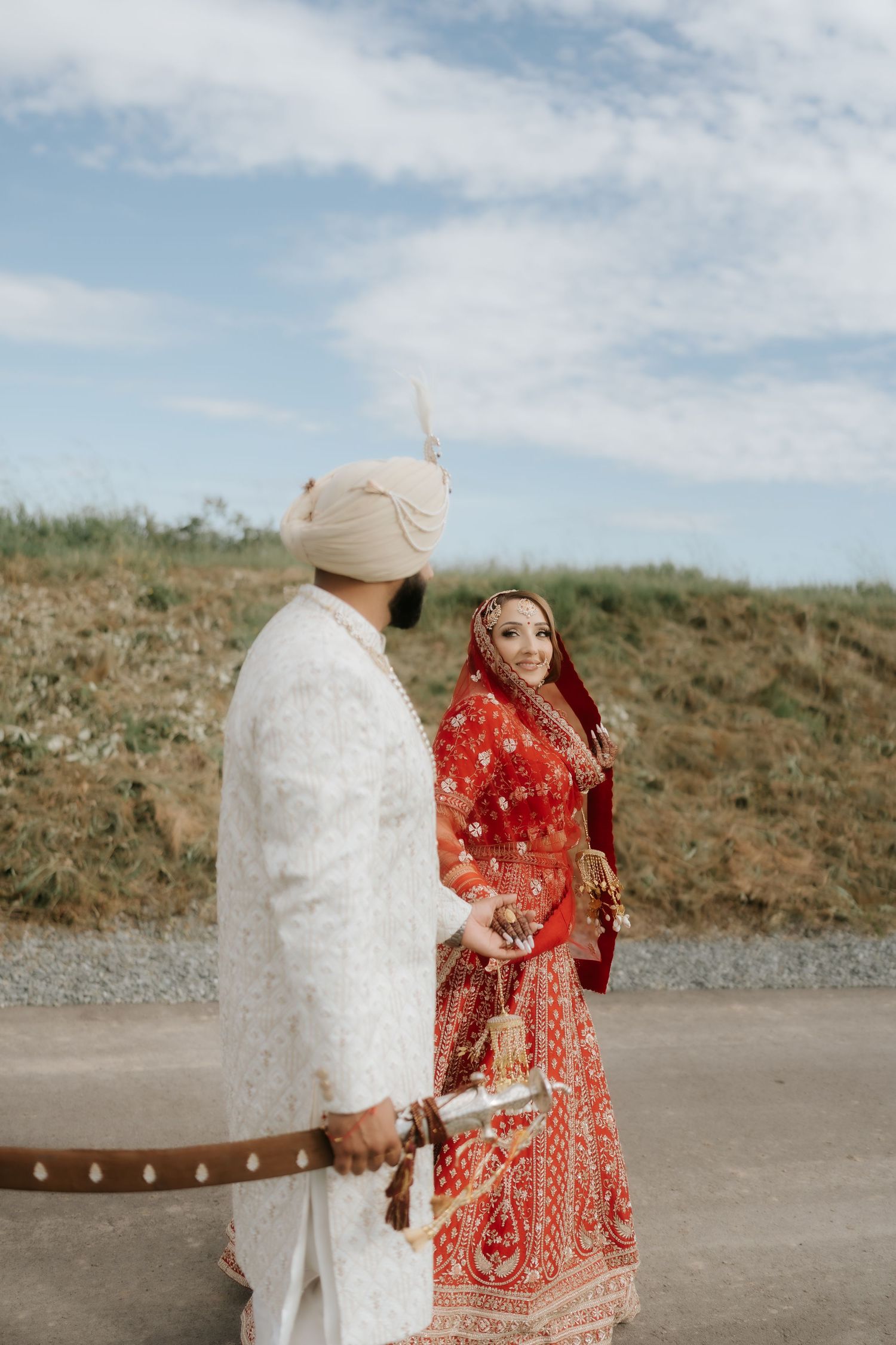 Sikh bride in red lehenga and groom in white sherwani with turban holding hands outdoors on their wedding day.