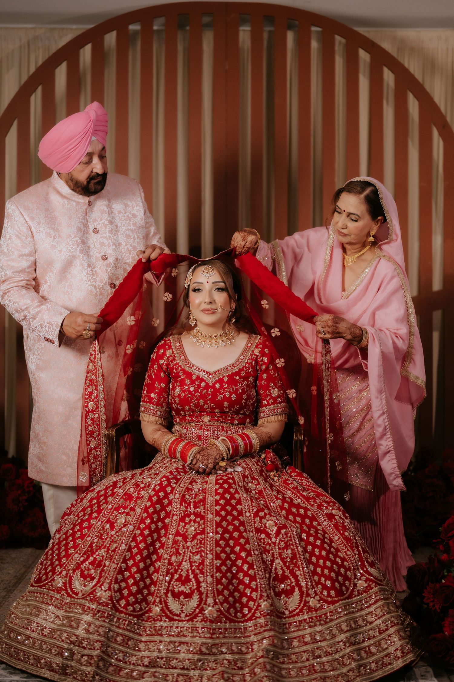 Sikh bride in red lehenga seated while parents drape her dupatta during traditional Indian wedding ceremony.