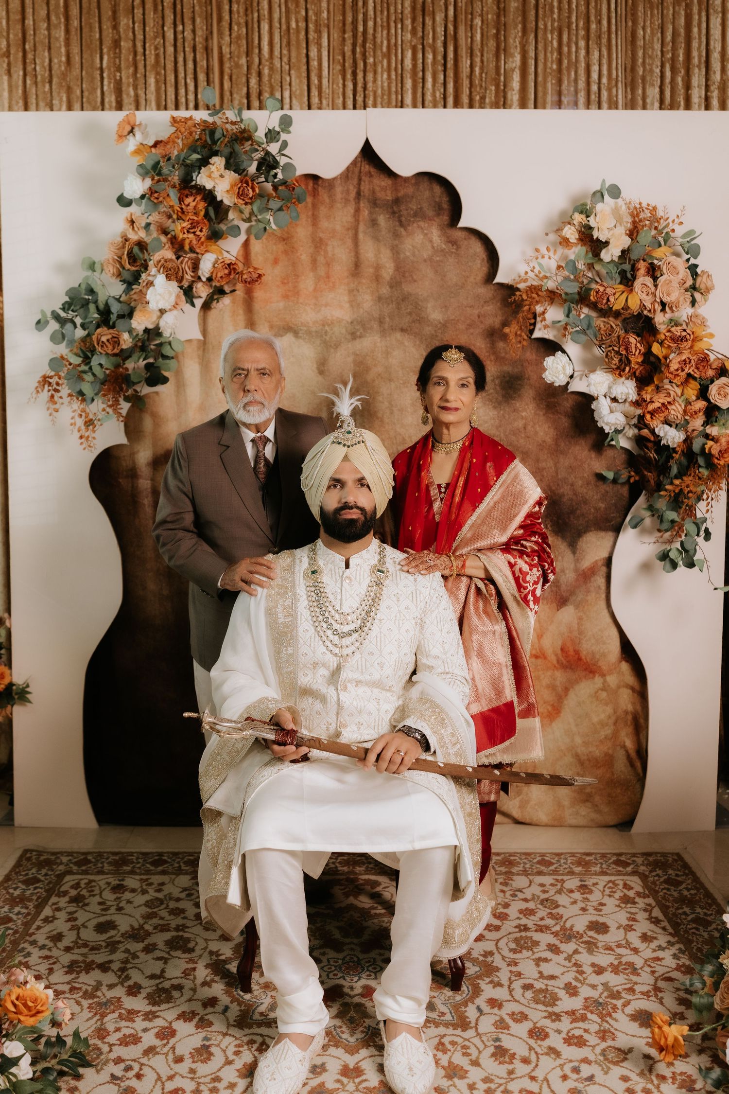 Sikh groom in white sherwani and turban seated with parents before floral backdrop at traditional Indian wedding.