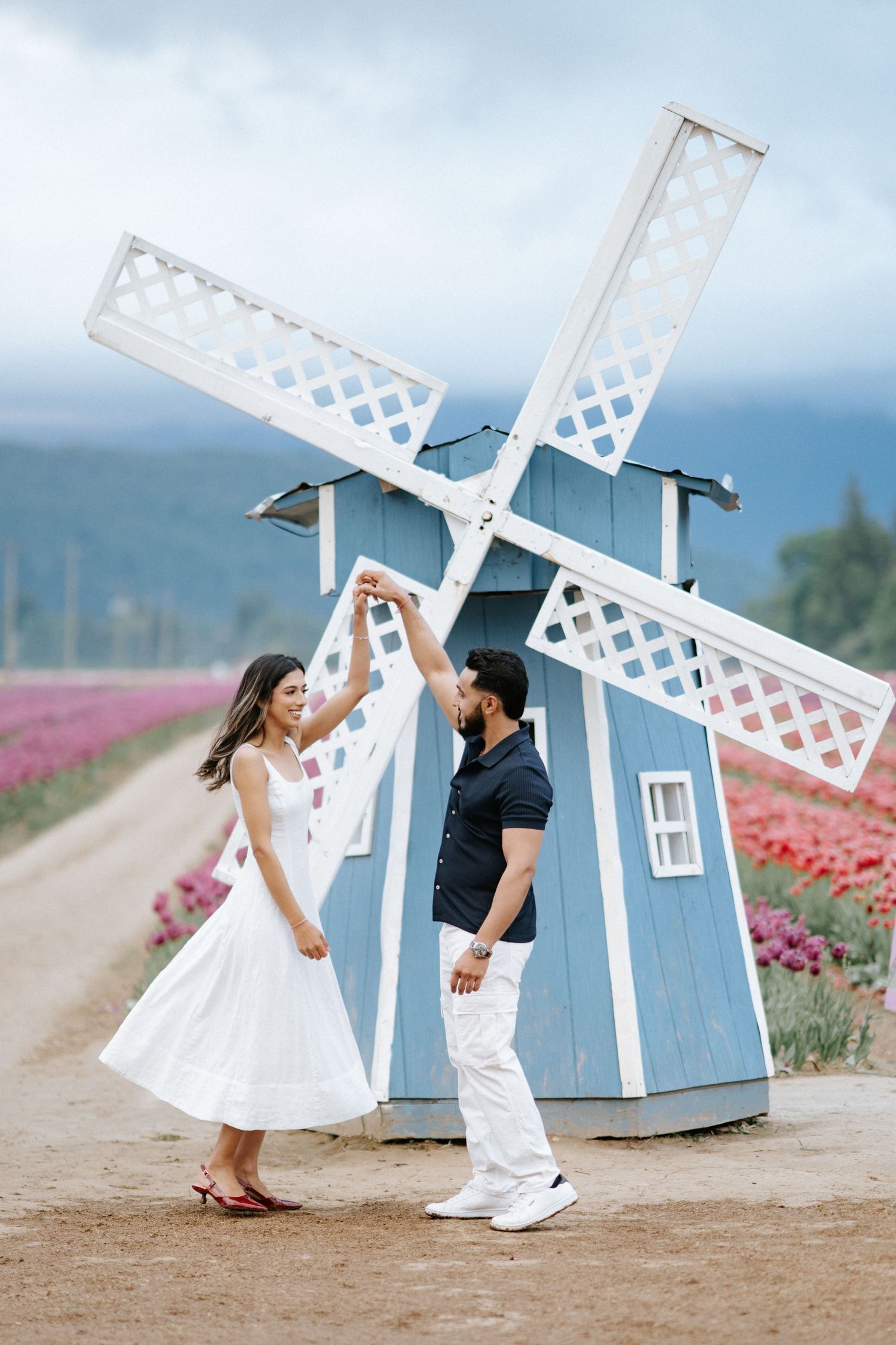A couple dances playfully in front of a blue and white decorative windmill in a flower field.