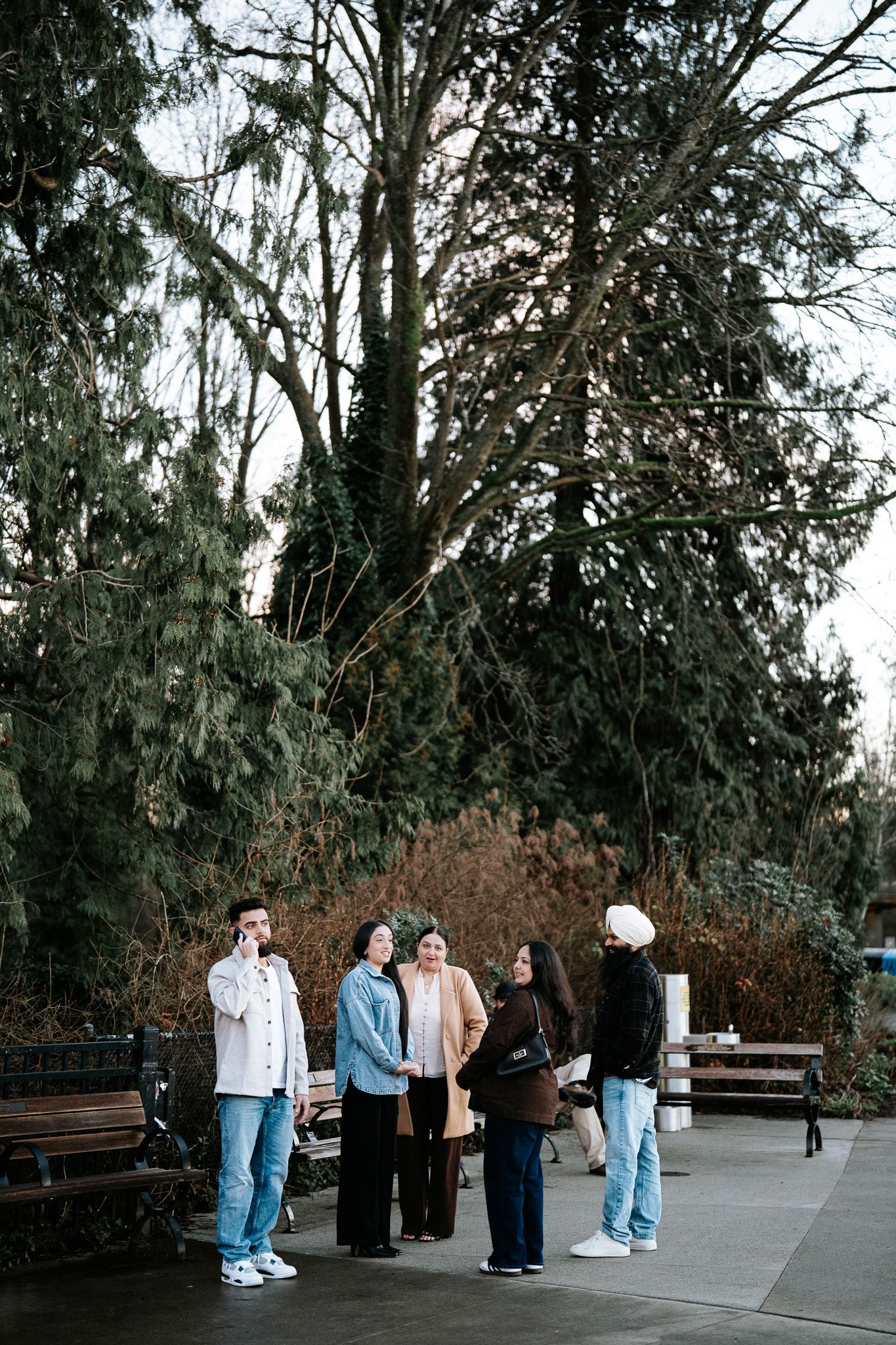 A sequence of candid outdoor photos showing a group gathering under tall evergreen trees in a park setting.