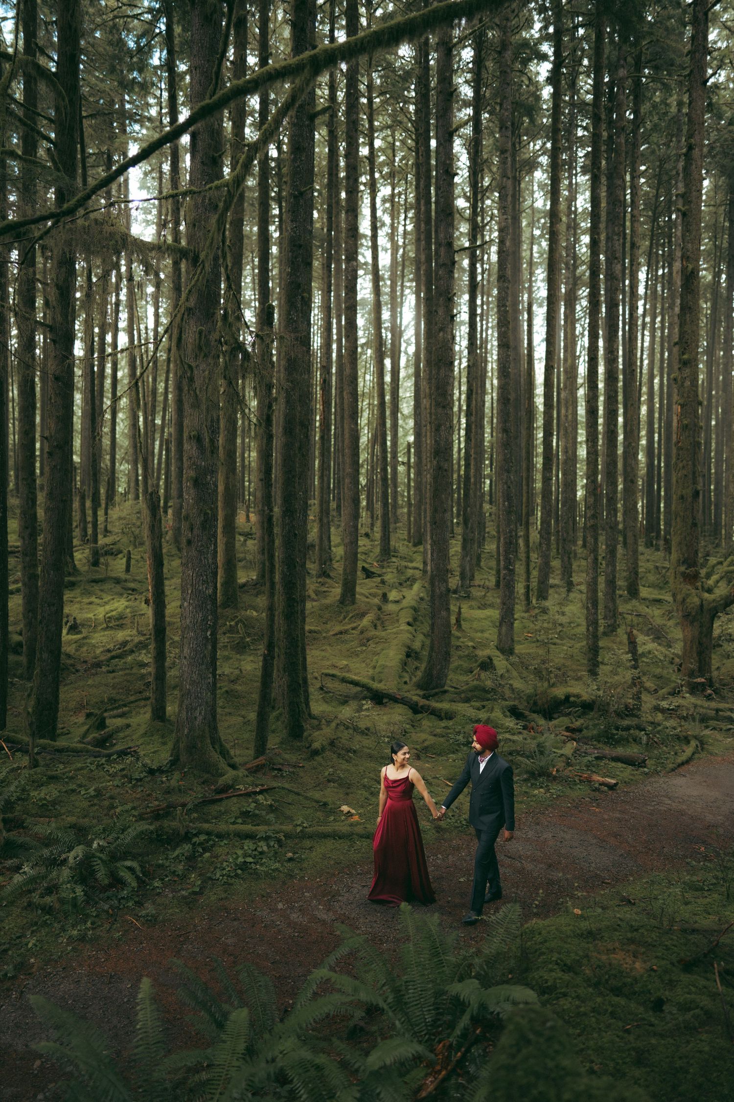 Two silhouettes walk along a winding forest path between towering pine trees.