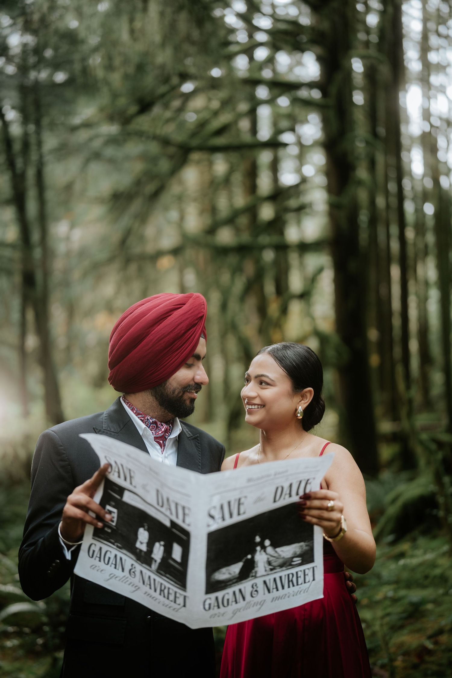 Couple shares a romantic moment while reading a newspaper-style save the date announcement in a forest setting.