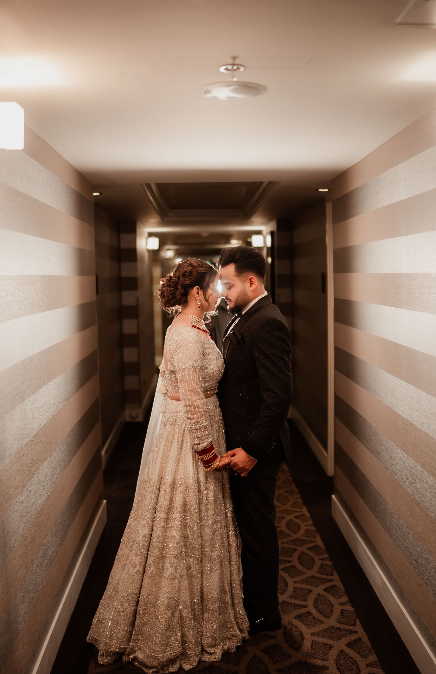 A romantic moment captured in a dimly lit hotel hallway with a couple in traditional South Asian wedding attire.