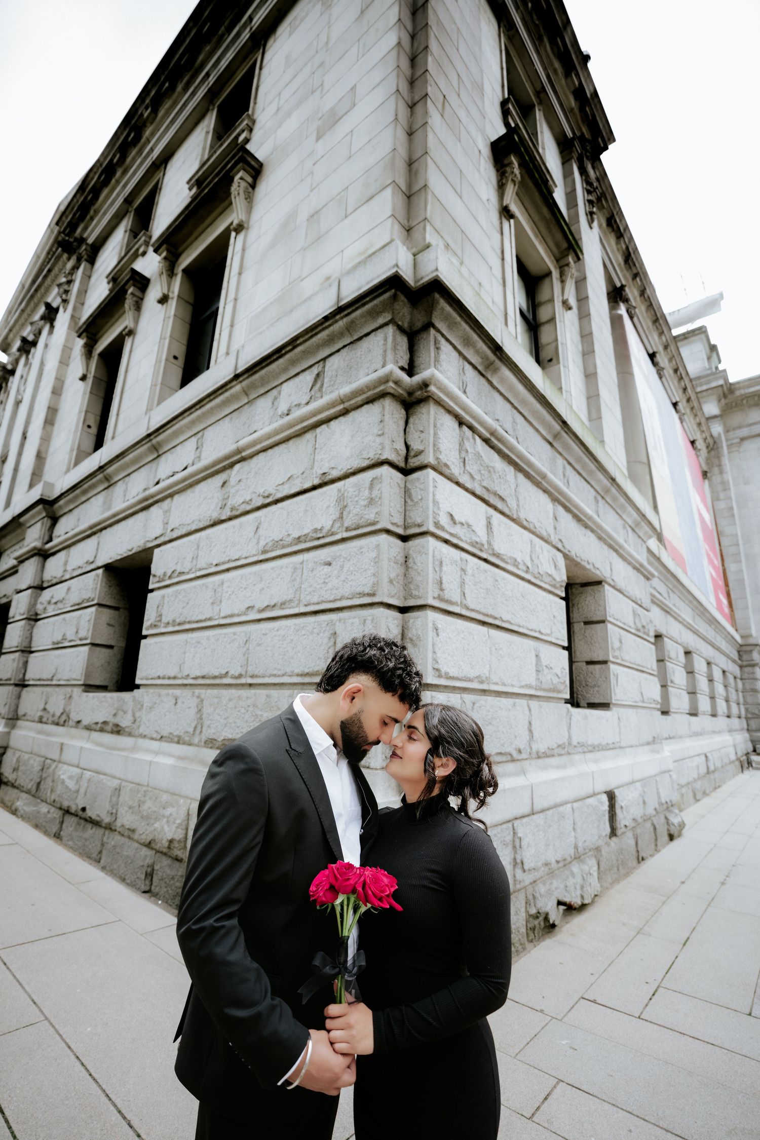 Couple in black attire shares tender moments with red flowers against stone building.