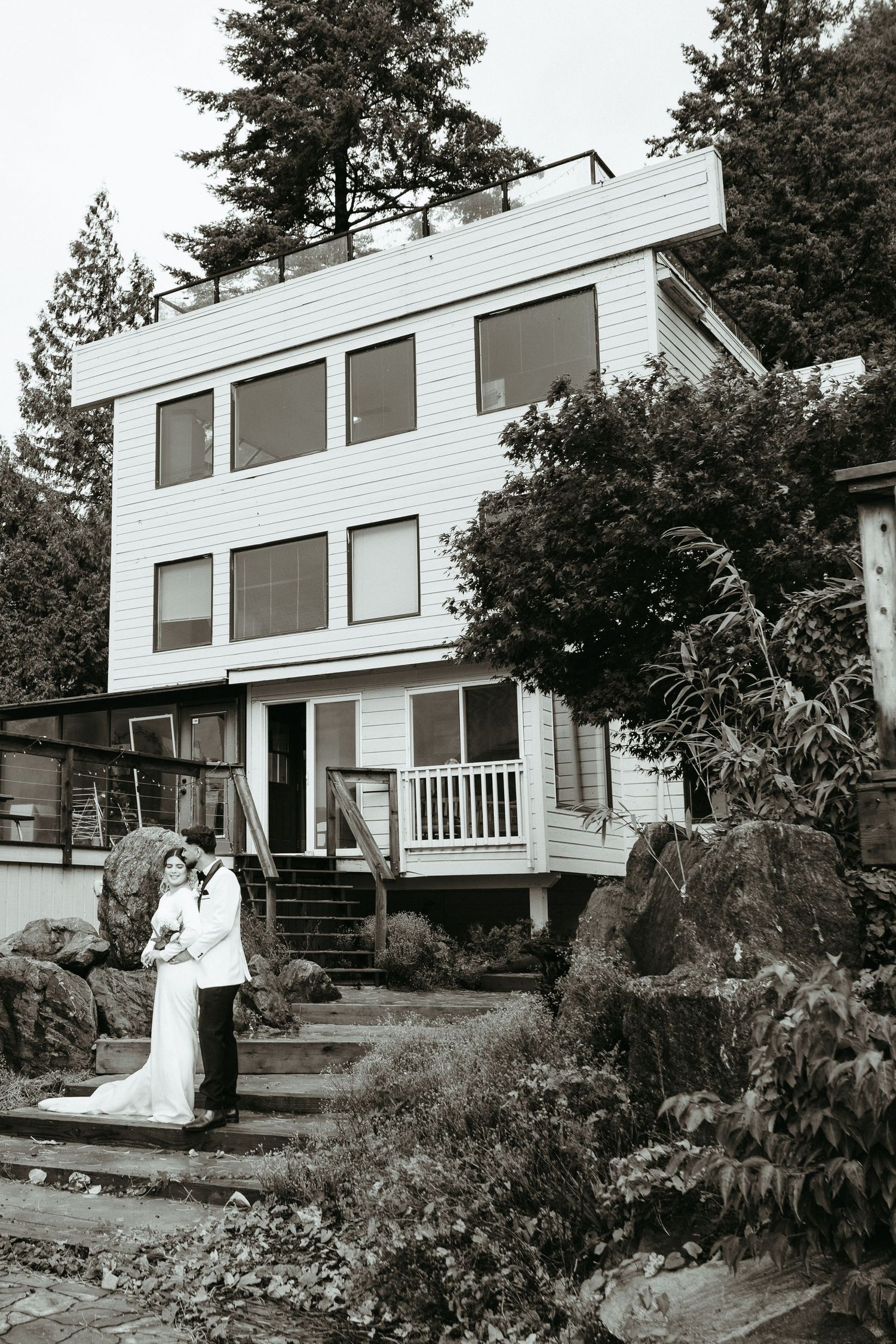 Modern white residential house with large windows set against wooded hillside landscape showing multiple exterior angles.